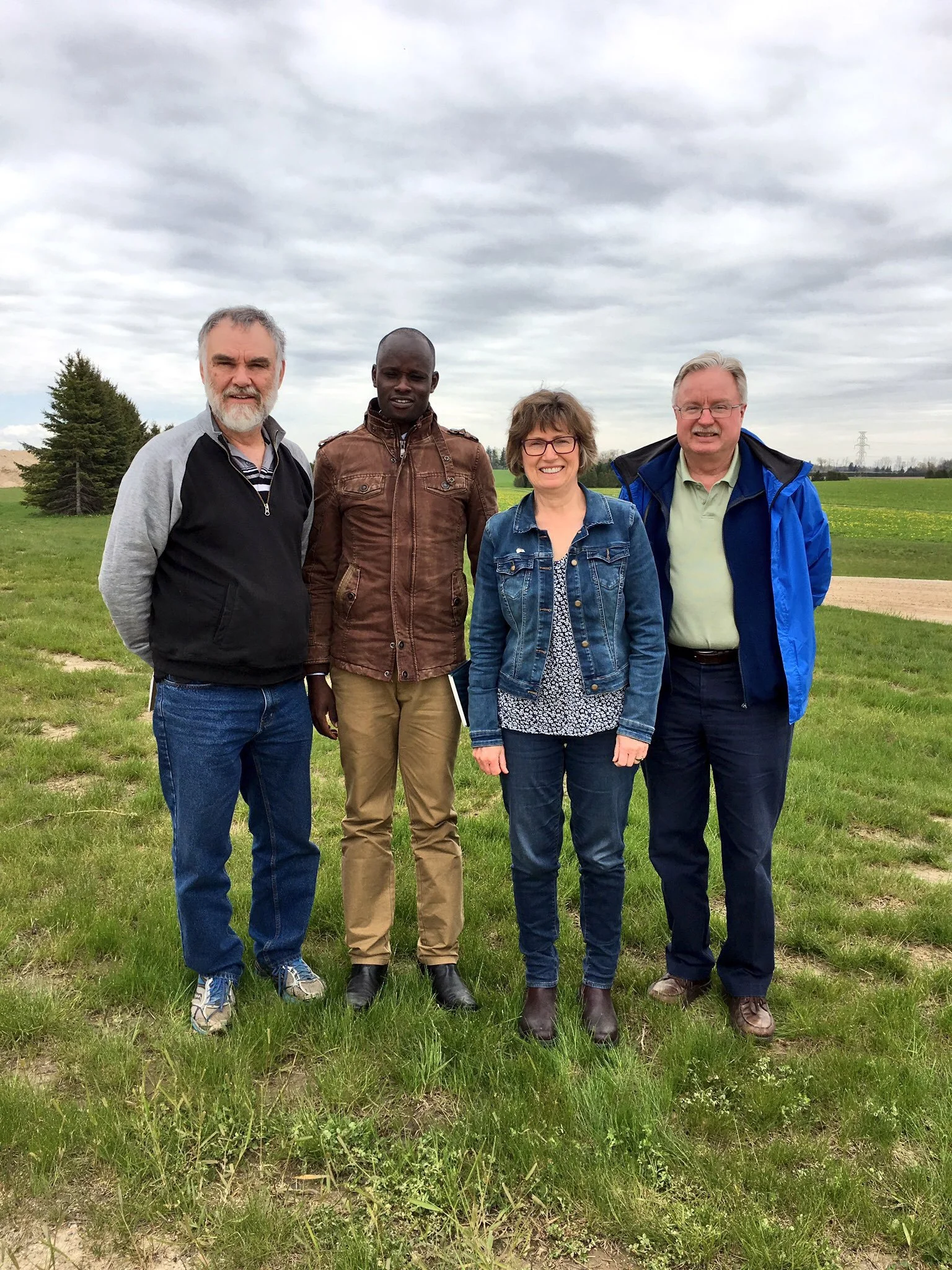 Four people standing outdoors on grass with cloudy sky and trees in the background.