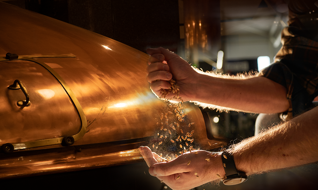 A person handling grains over a copper kettle in a brewery