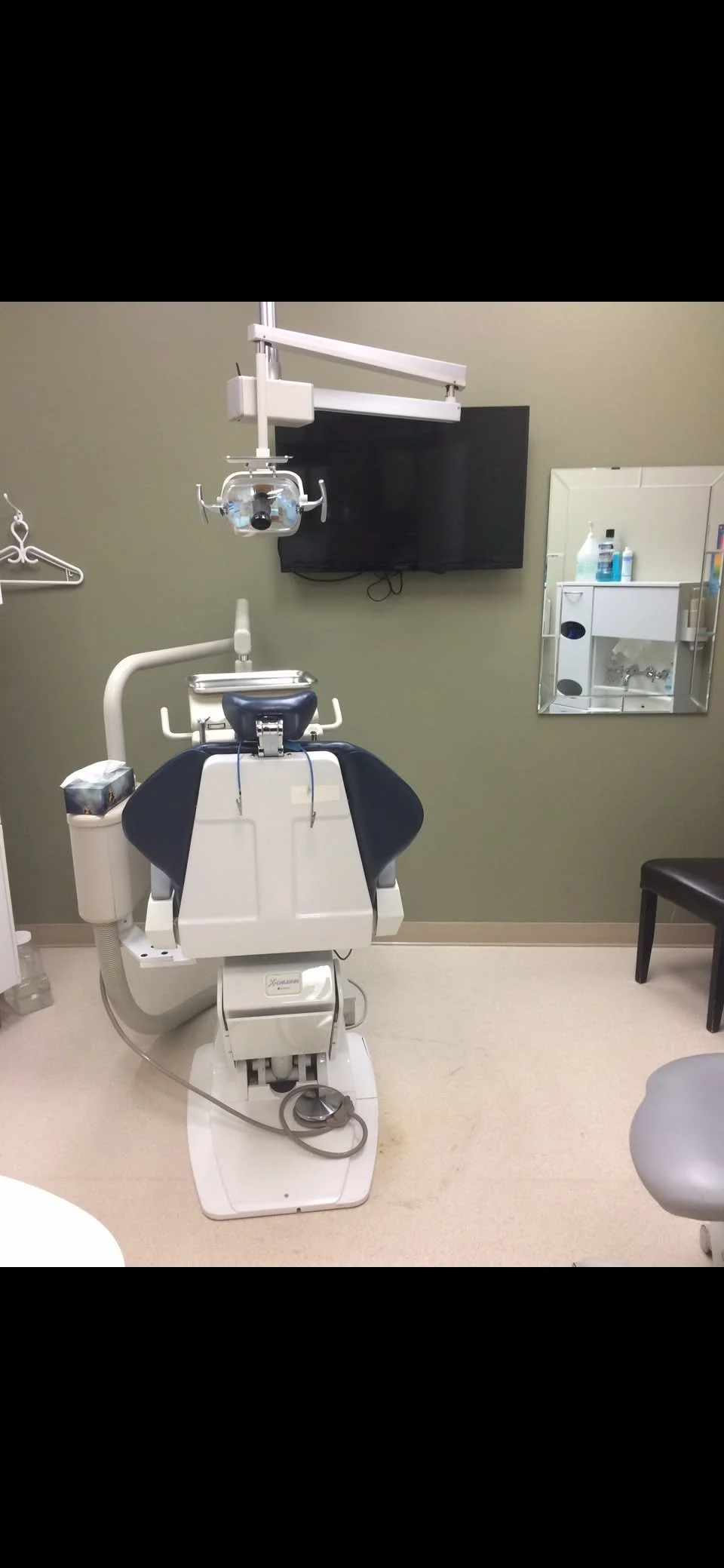 Dental exam room with dental chair, overhead light, monitor, mirror, stool, and dental supplies.
