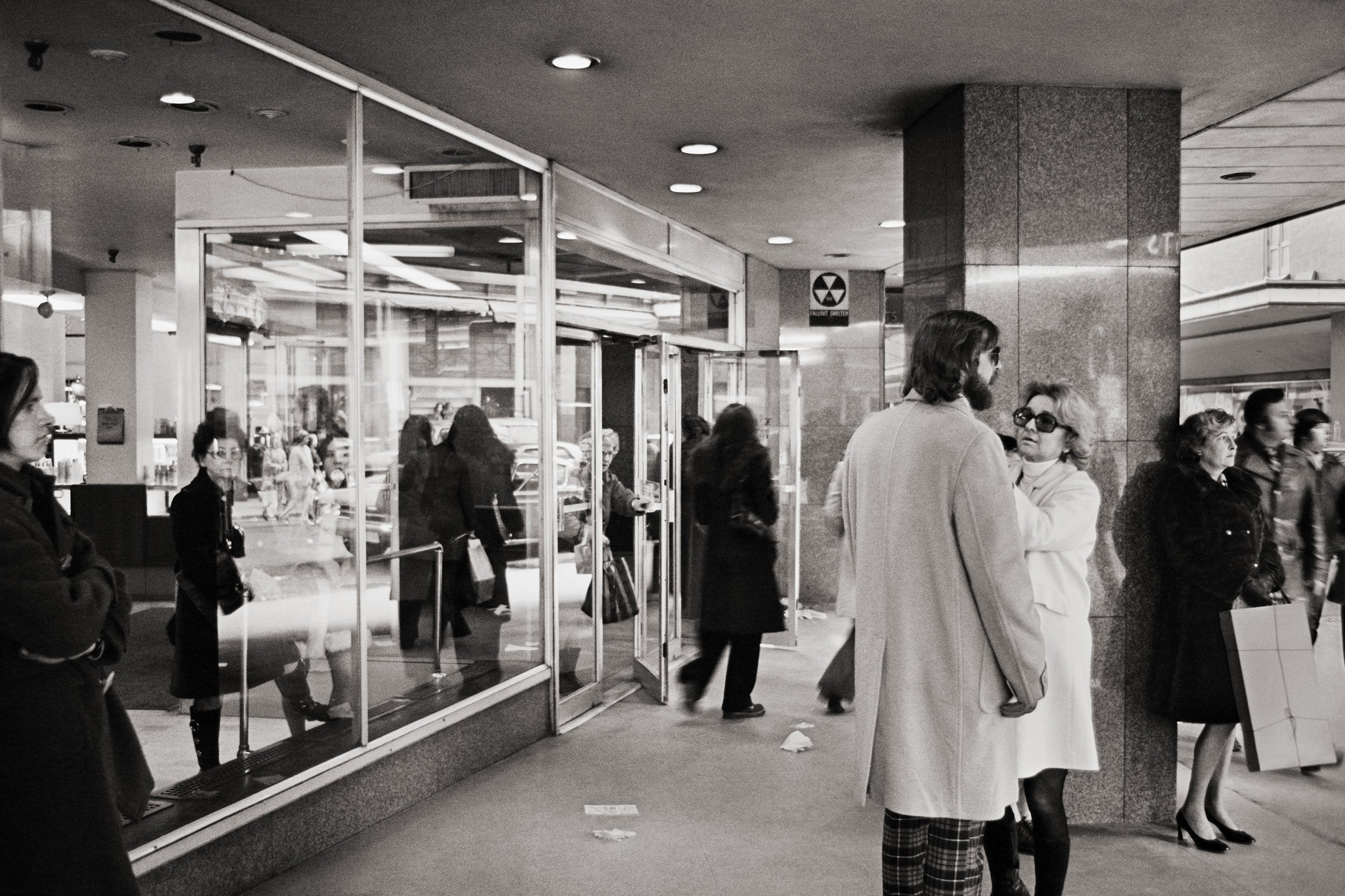 People waiting in line inside a busy transportation terminal, with some people standing near glass barriers and others walking, and a sign indicating a bomb threat warning.