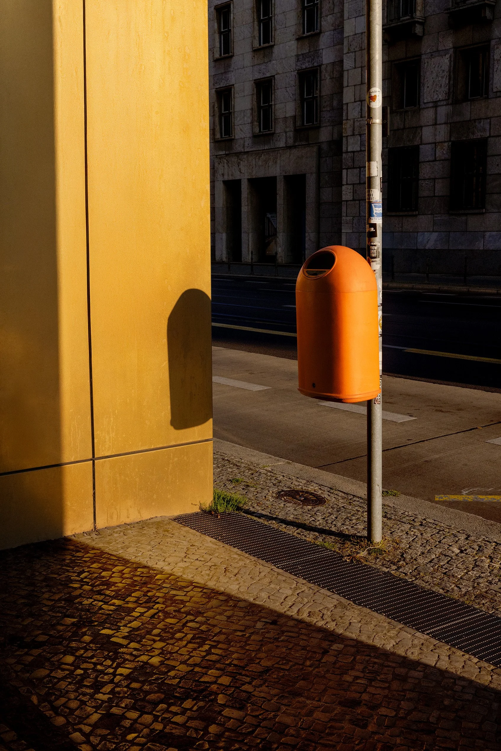 Street scene at night showing a yellow building wall on the left, a cobblestone sidewalk, a bright orange trash bin, and a street pole covered with stickers, with a stone building in the background.