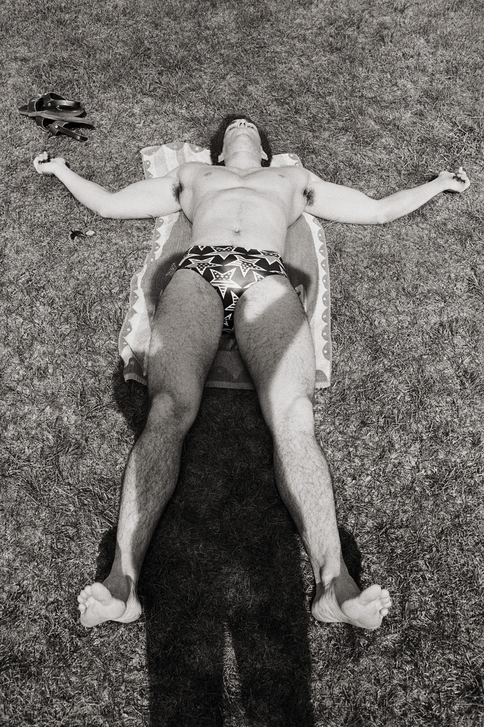 A man lying shirtless on the grass with arms and legs spread out, wearing patterned shorts, next to flip-flops.