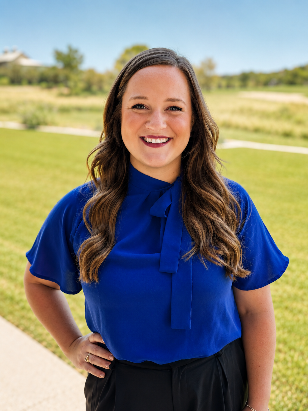 A woman with long wavy brown hair, wearing a bright blue blouse with a bow tie at the collar, standing outdoors on a sunny day with green grass and trees in the background, smiling at the camera.