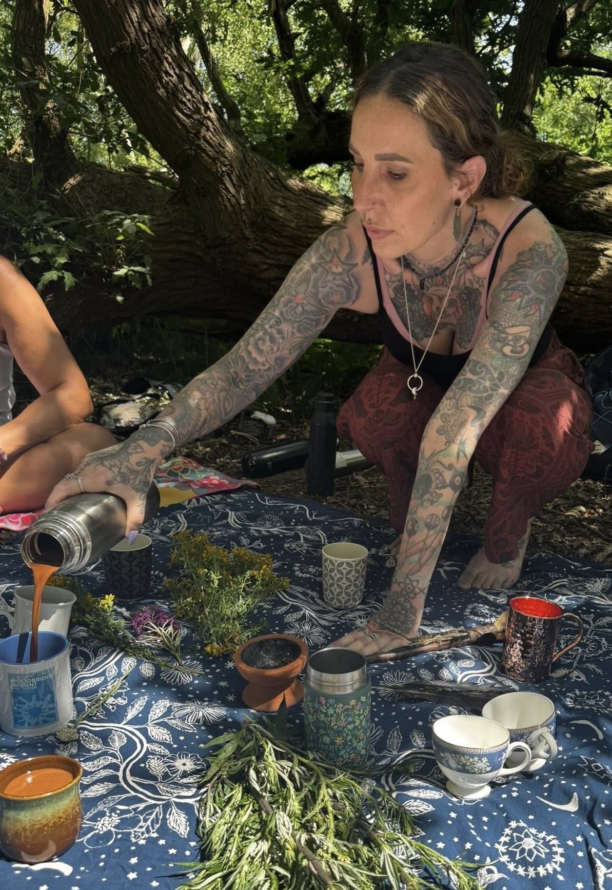 A woman with tattoos on her arms and shoulders, wearing a tank top and red patterned pants, is pouring a drink from a stainless steel thermos onto cups on a table. She is outdoors under a large tree with green foliage, with a patterned tablecloth, cups, and plants on the table.