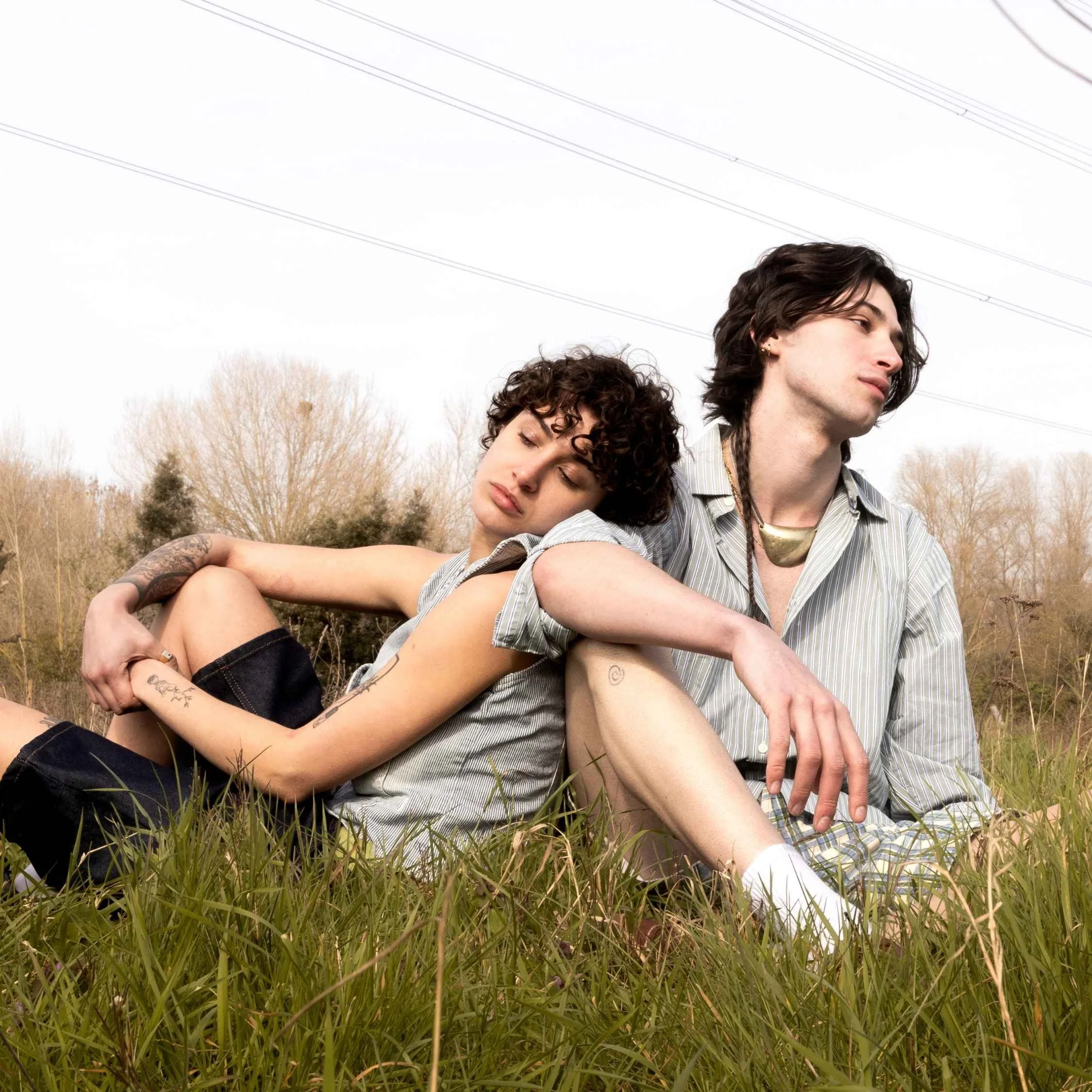 Two young people, a woman with curly hair and a man with long dark hair, sitting in a grassy outdoor area during the daytime. The woman rests her head on the man's shoulder with her eyes closed, and the man looks into the distance. The background fea