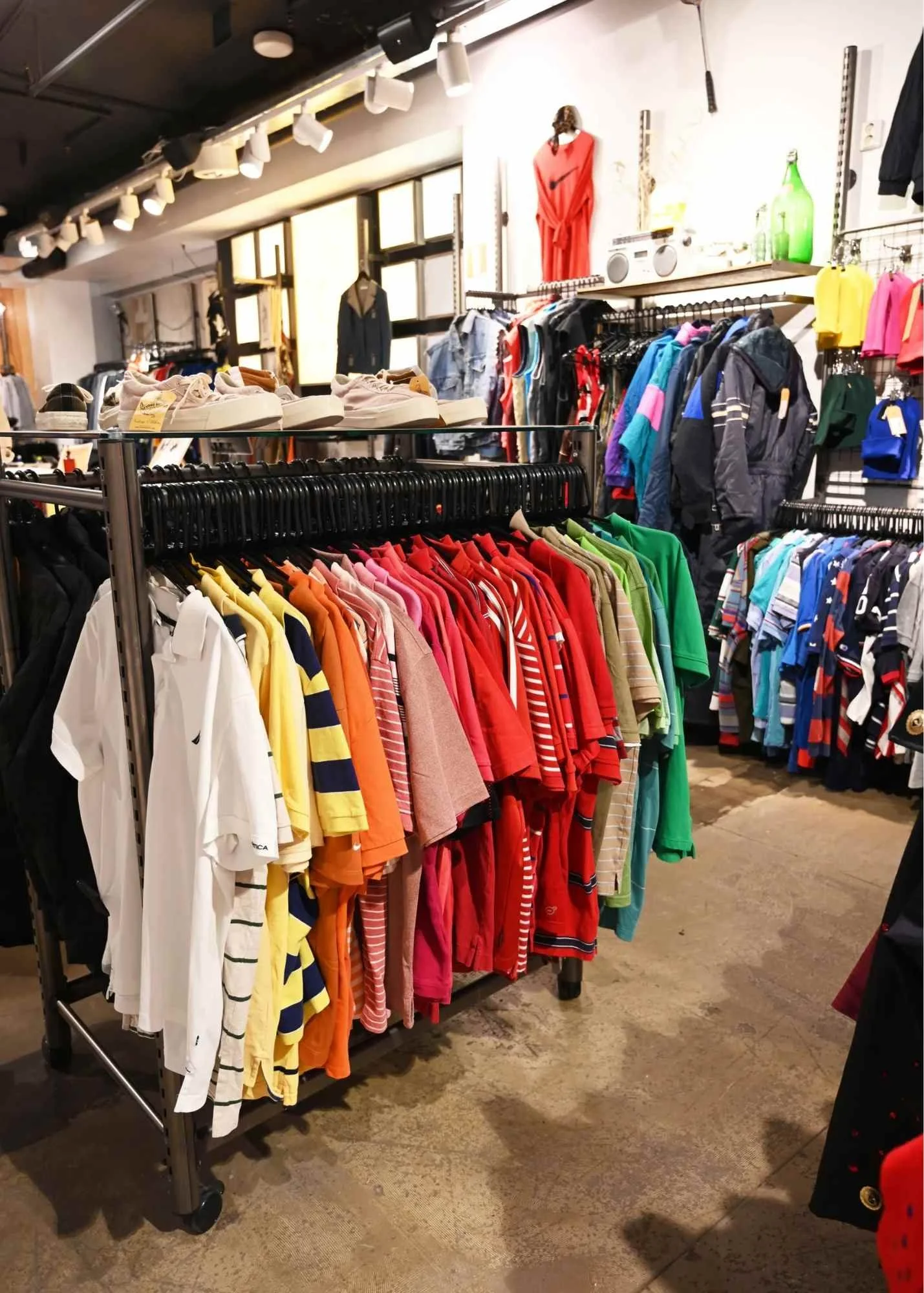 Clothing store with colorful shirts and jackets on racks, shelves with shoes, and decorative items like a dress on a mannequin and green bottles on a shelf.
