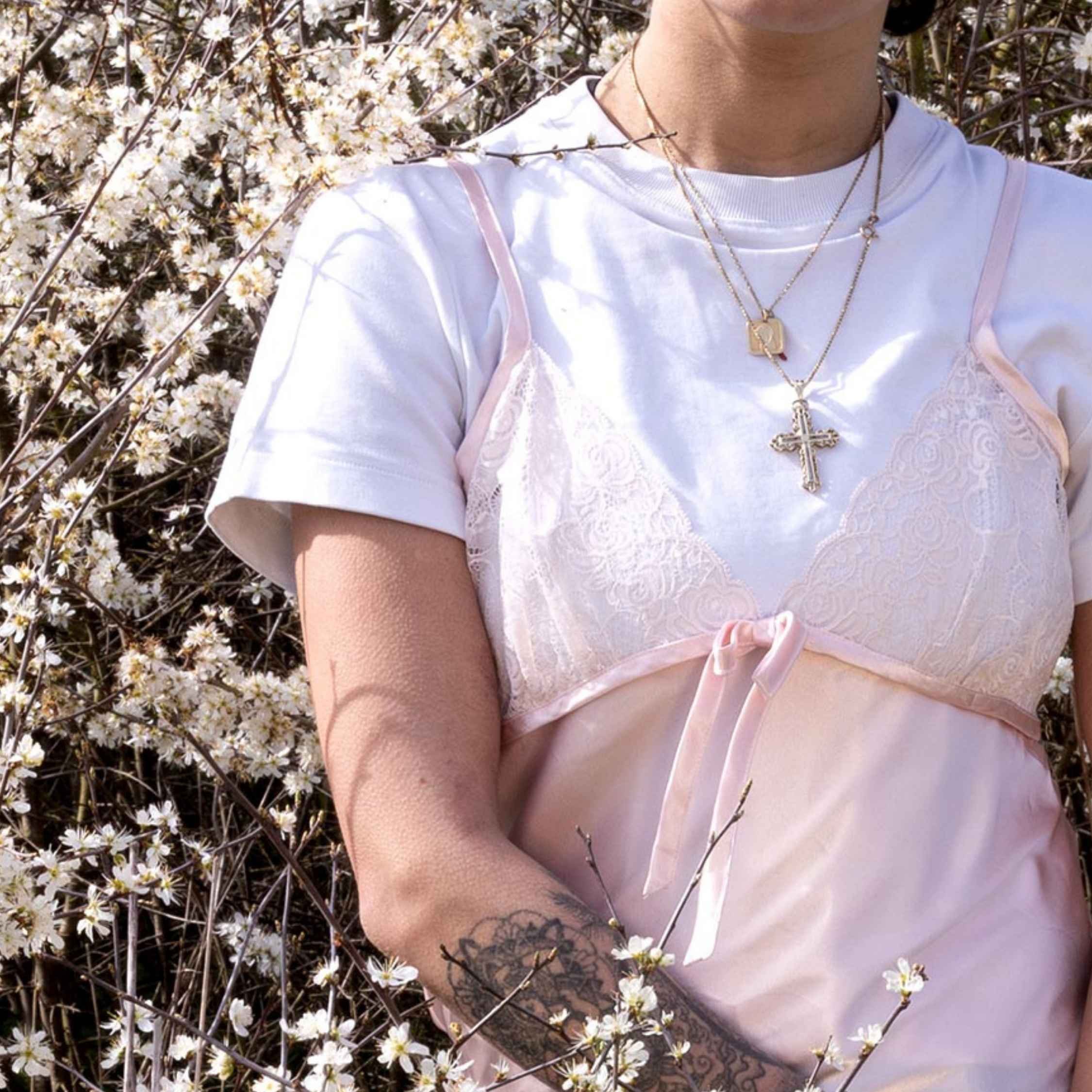 A person wearing a white top with pink accents and gold necklaces with cross pendants, standing among white flowering branches.