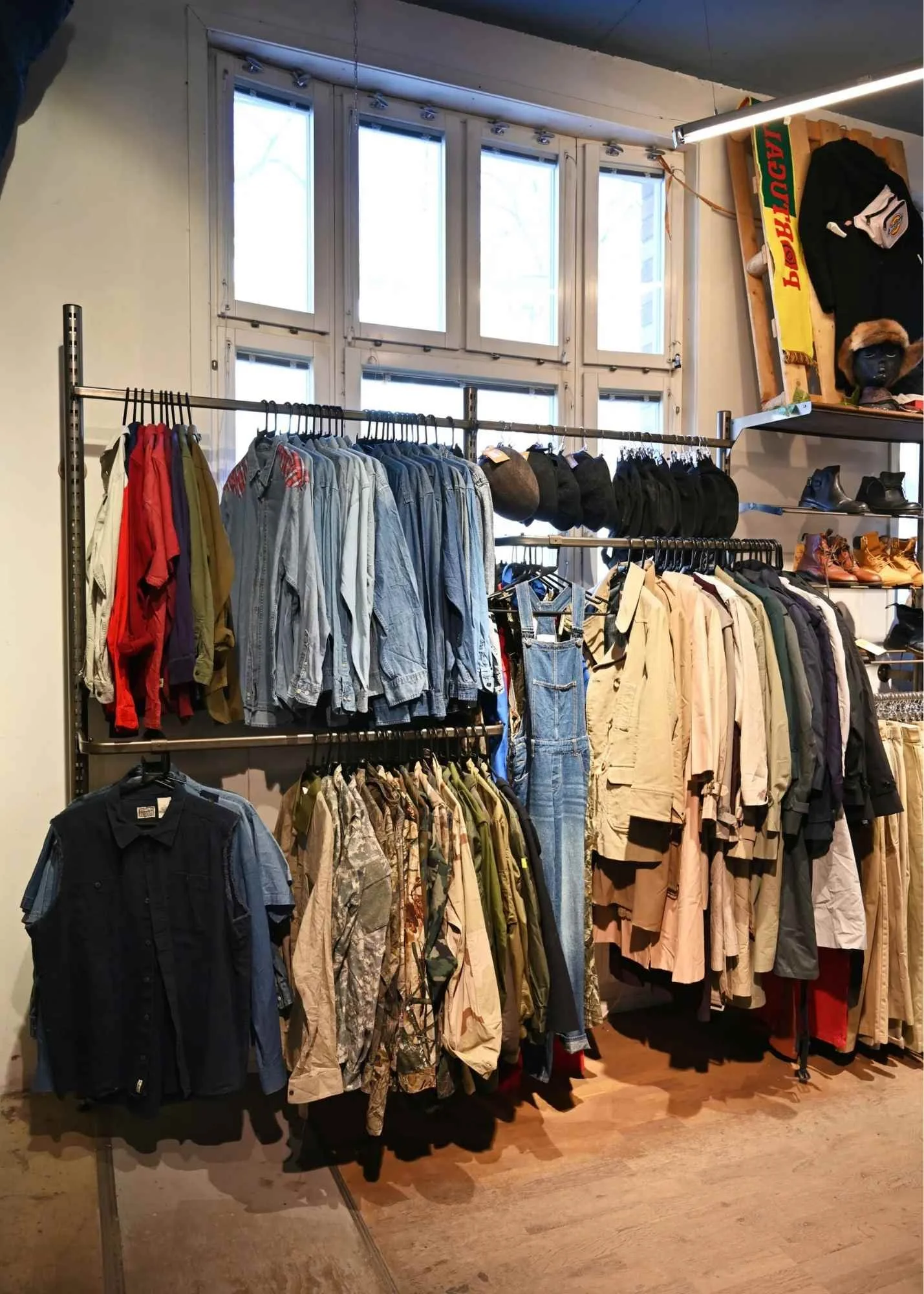 Clothing display in a store with shirts, jackets, and hats on metal racks, and a wooden shelf with boots and accessories, near windows.