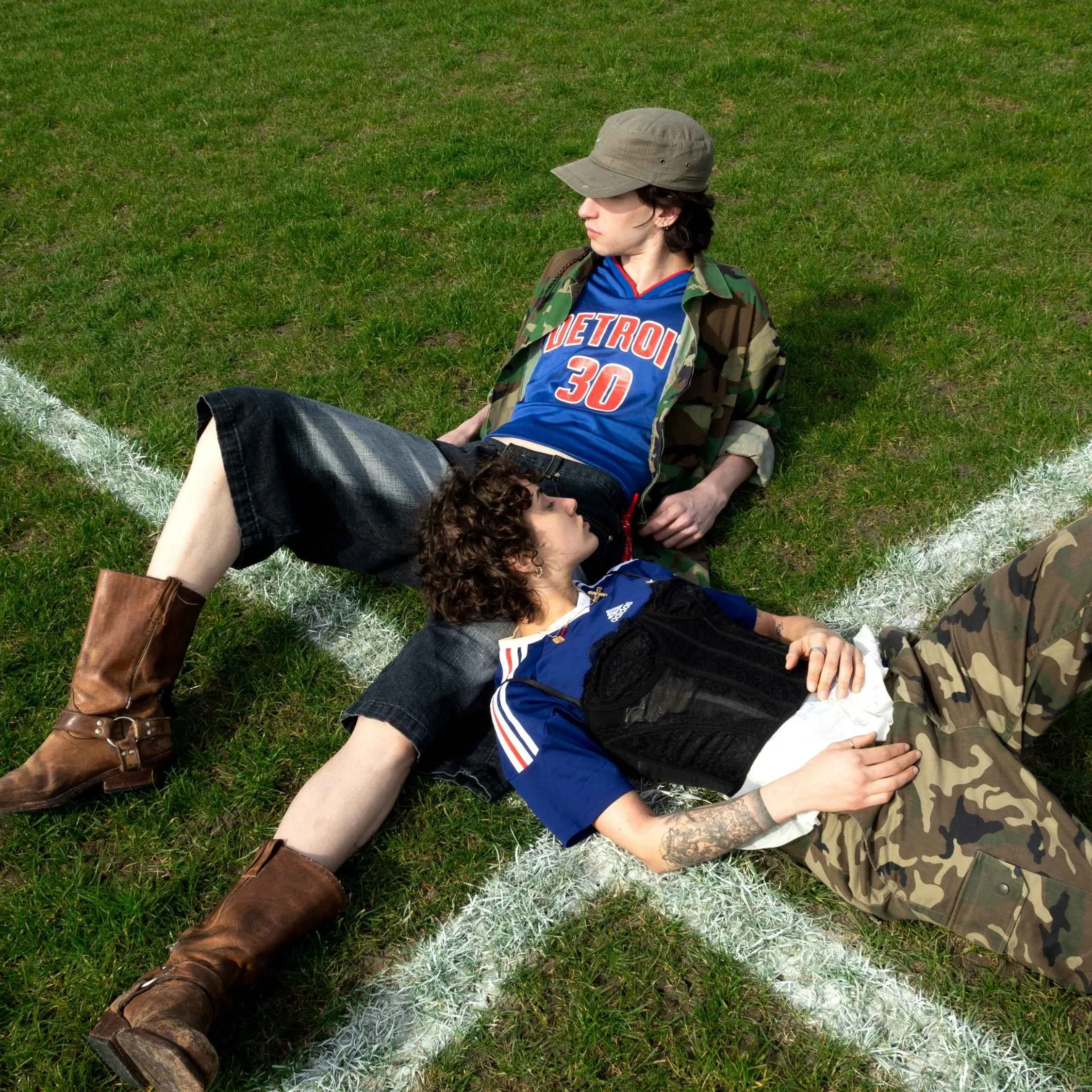 A person with shoulder-length dark hair wearing a gray hat, a Detroit basketball jersey numbered 30, a camouflage jacket, and ripped jeans, sitting on grassy field with other person lying next to them.