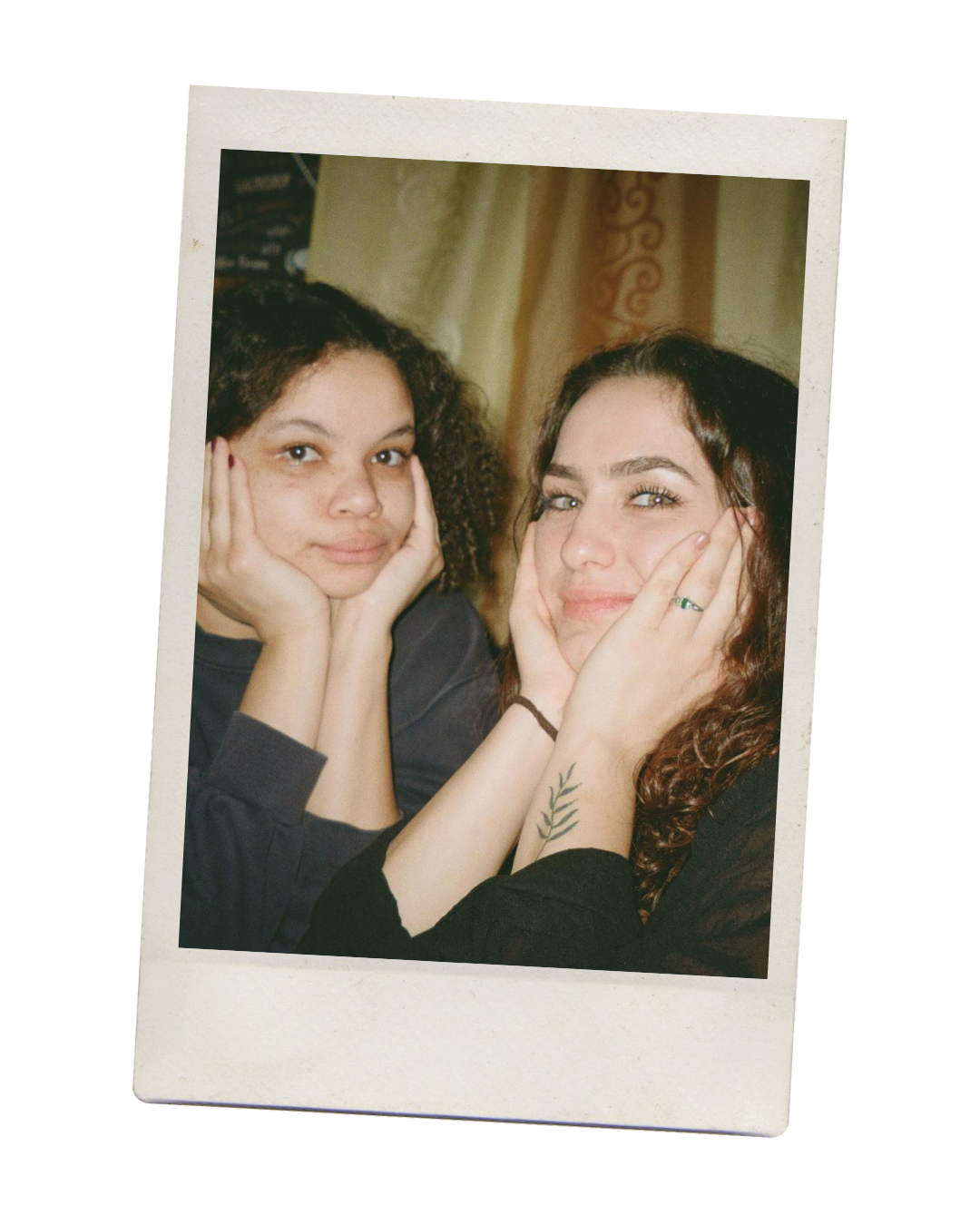 Two women with curly hair resting their cheeks on their hands, smiling at the camera, with a beige curtain in the background.