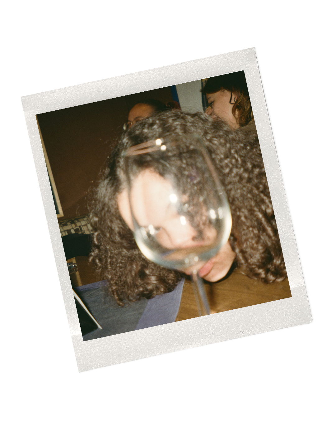 Person with curly hair leaning over a table with a glass of water in the foreground, with two women in the background smiling and engaging in conversation.