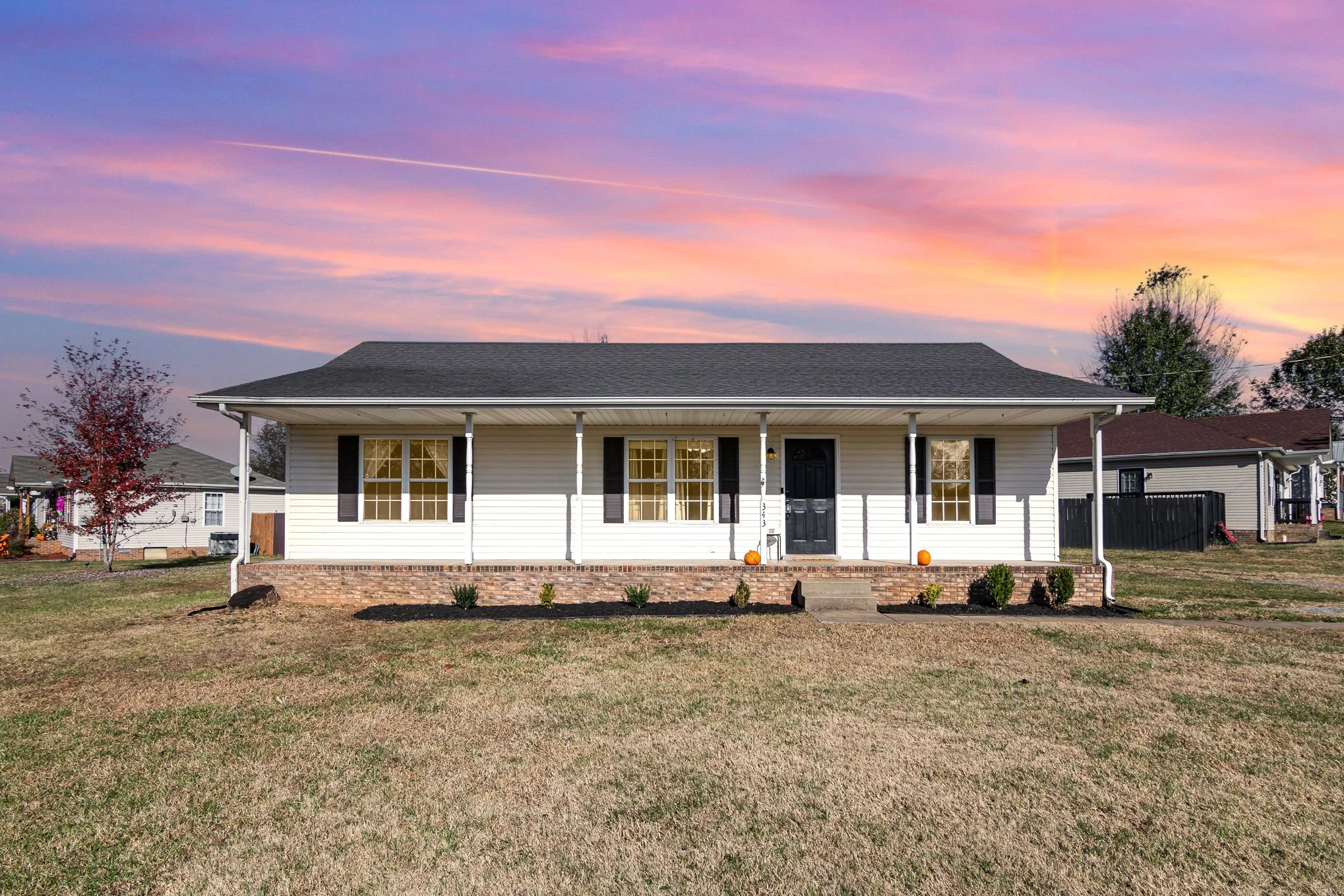 Front view of a single-story house with white siding, black front door, and two large windows, decorated with pumpkins outside; there are neighboring houses and trees, with a colorful sunset sky in the background.