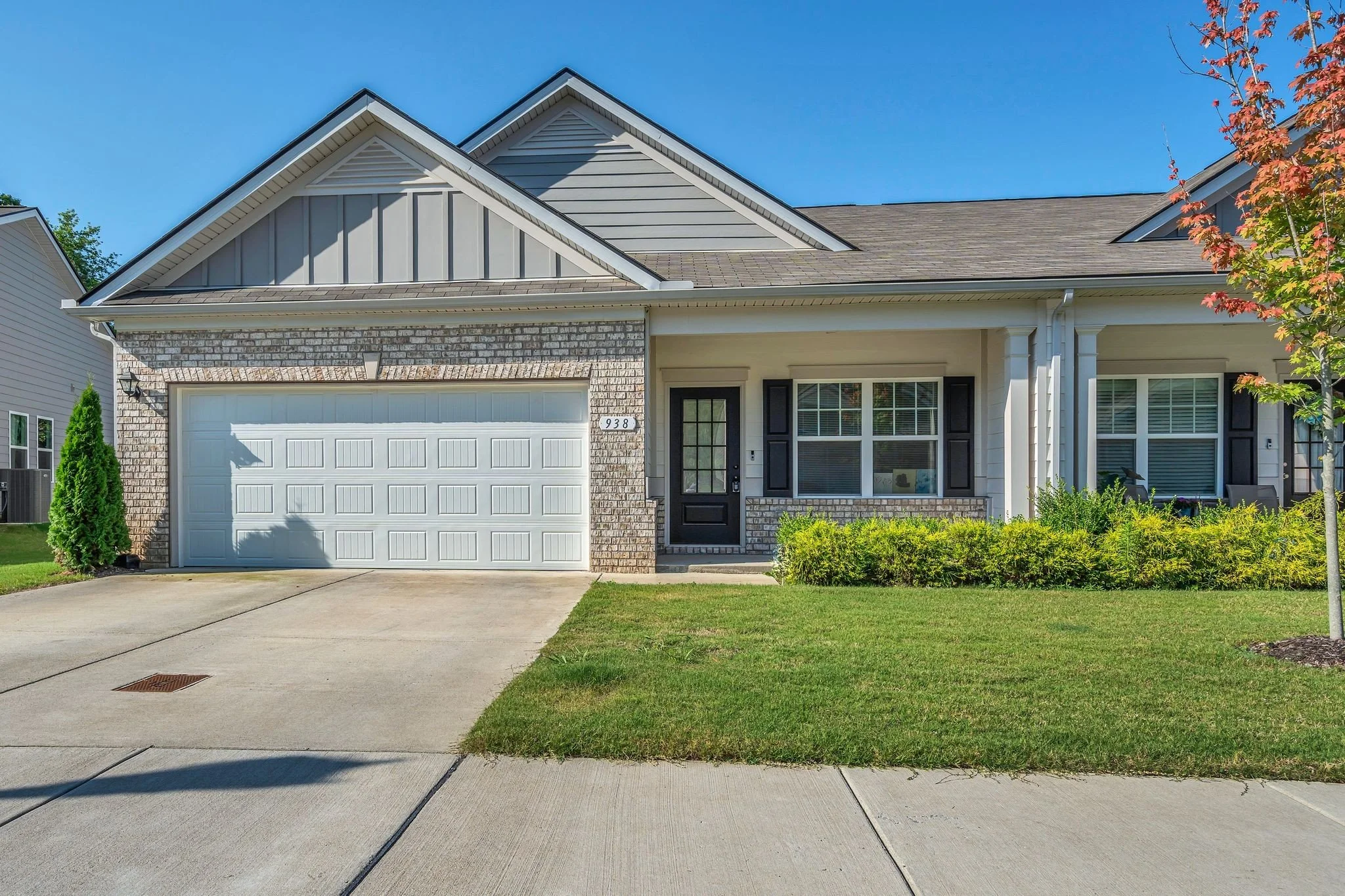 Front view of a suburban house with a brick and siding exterior, a white garage door, black front door, and some greenery and trees in the yard under a clear blue sky.