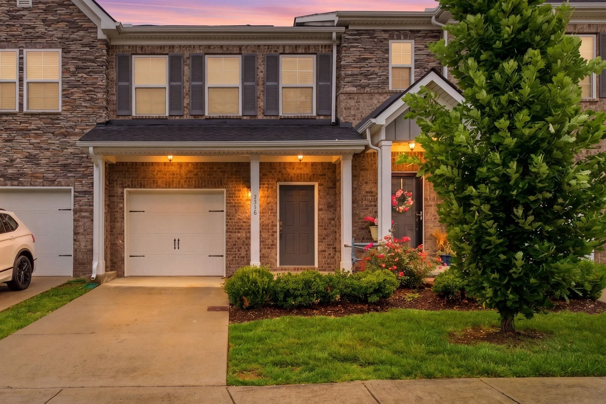 A brick townhouse with two garage doors, a dark front door with a wreath, and a porch with two columns. There are windows with shutters on the second floor, a tree in front, and flowers and bushes near the entrance. The sky is pink and purple during sunset.