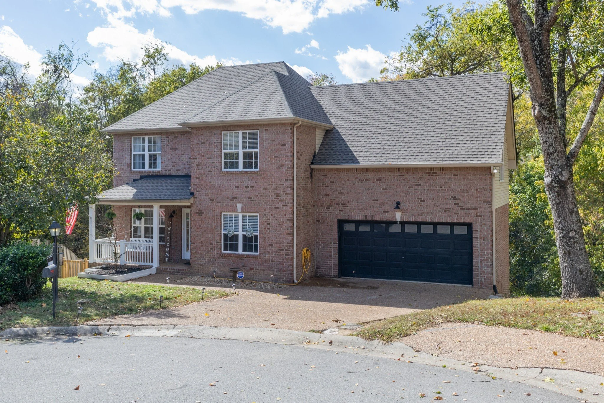 A two-story brick house with a black garage door, large windows, and a small front porch with white railings, surrounded by trees and grass, under a partly cloudy sky.