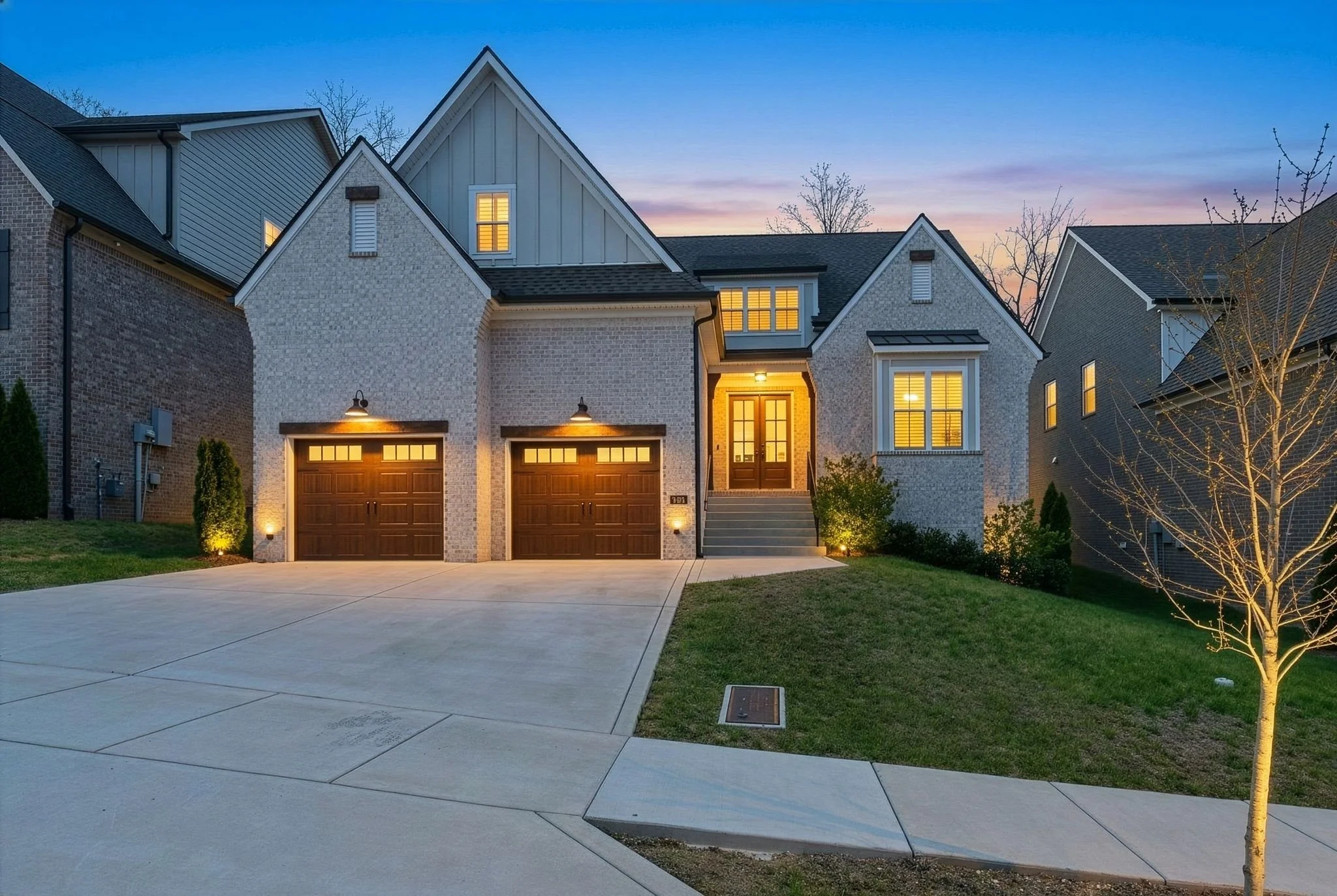 A modern suburban house at dusk with illuminated windows, a double garage with wooden doors, a staircase leading to the front door, and landscaped front yard with a small tree and outdoor lights.
