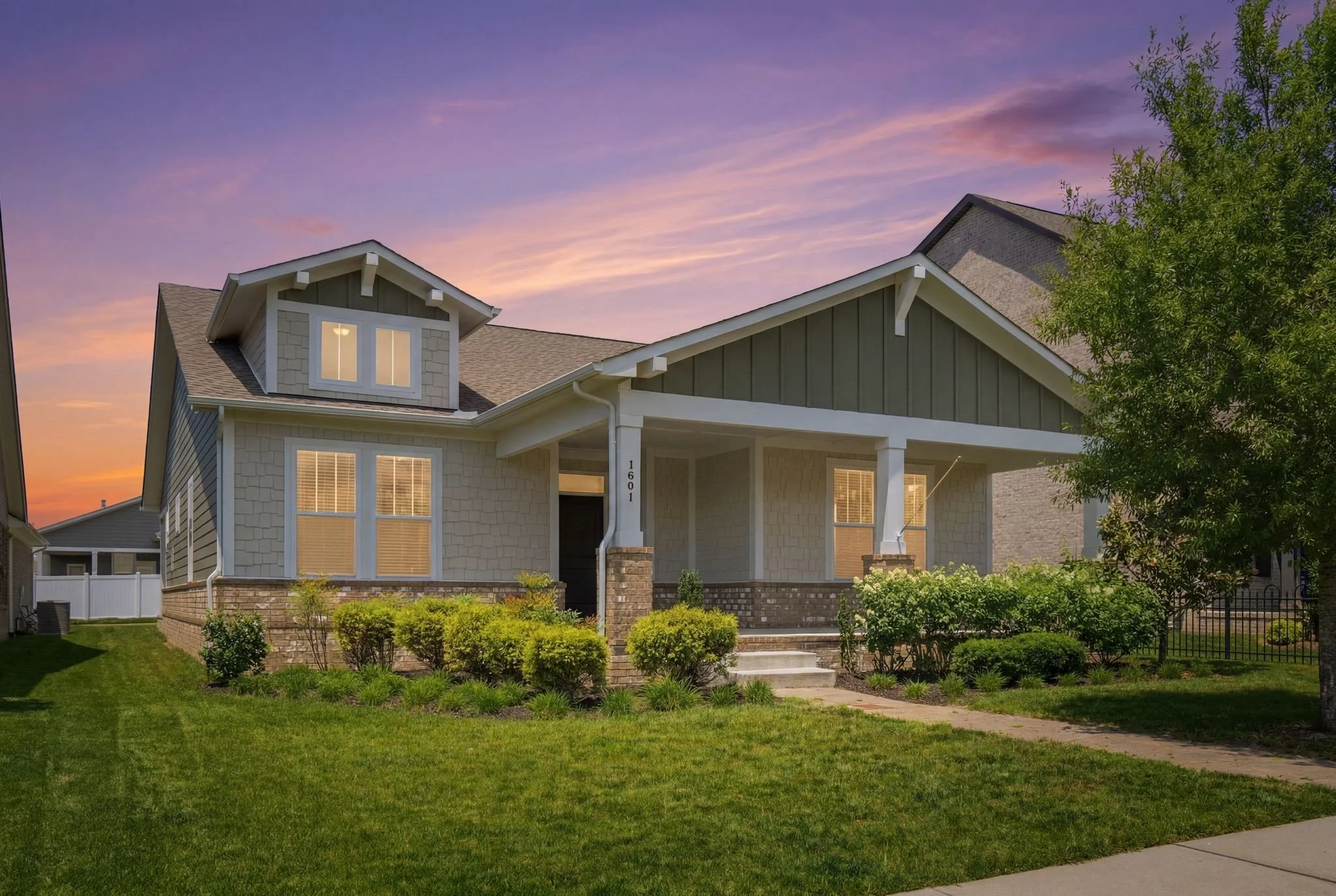 A two-story suburban house with a green exterior, tan shingles, and a front porch under a pink and purple sunset sky, surrounded by a well-maintained lawn and bushes.