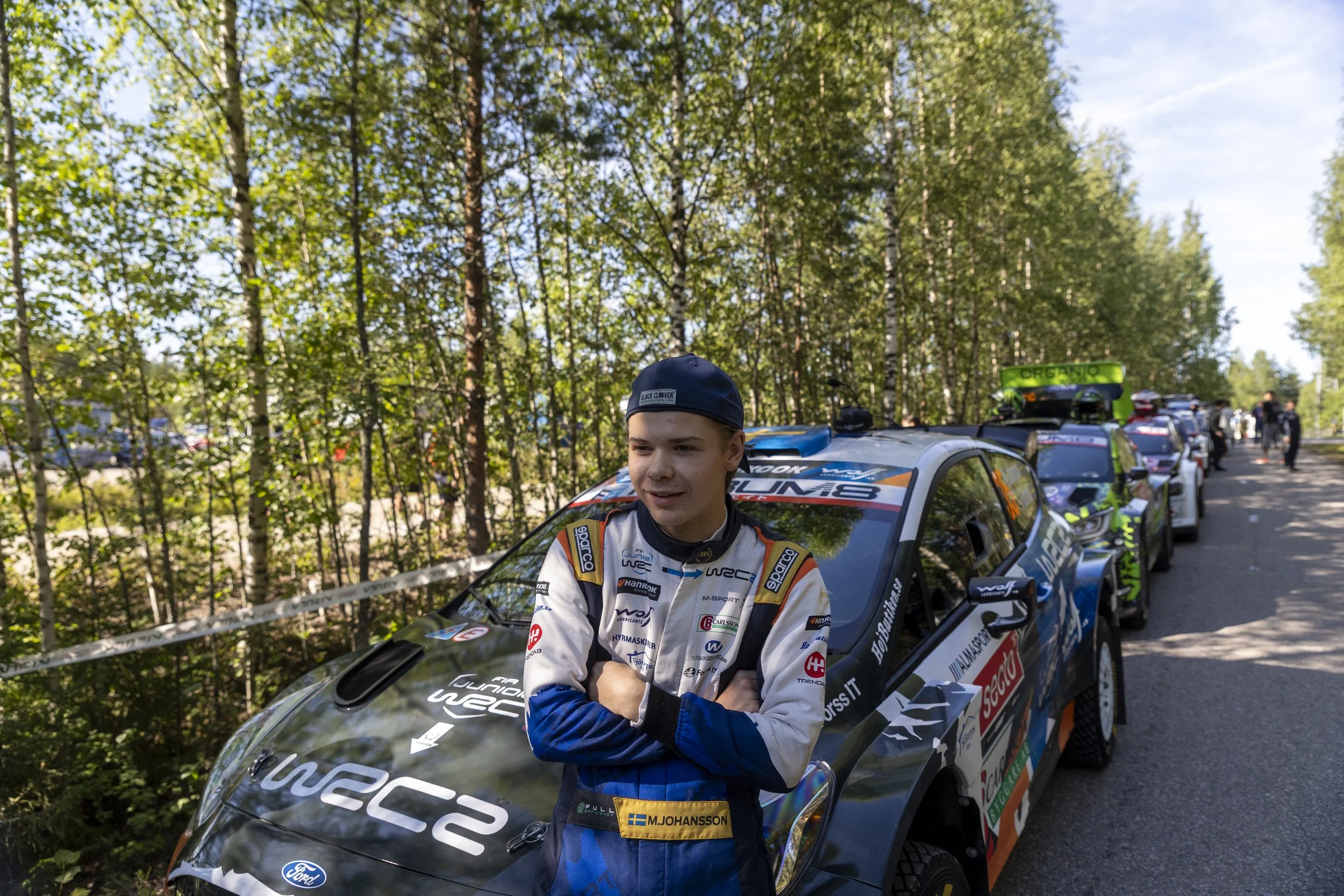 A young male rally car driver in a racing suit standing in front of a line of rally cars outdoors in a wooded area. He is wearing a dark cap with 