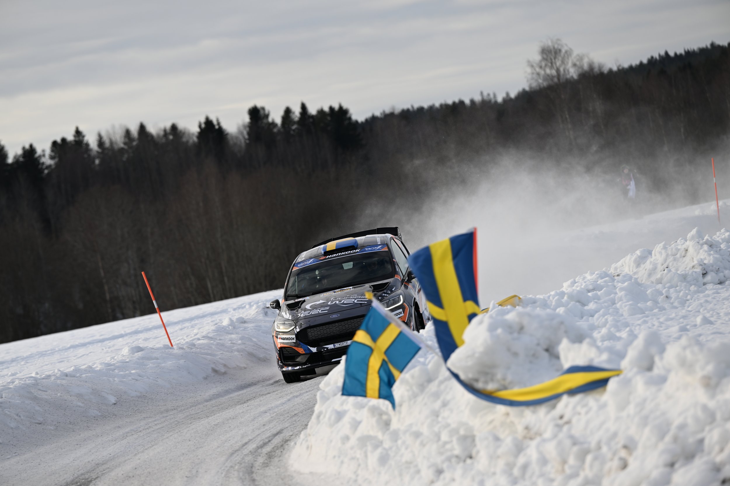 A race car speeds through a snowy landscape, creating a trail of snow and ice behind it, with Swedish flags and orange poles marking the course, set against a backdrop of trees and a cloudy sky.