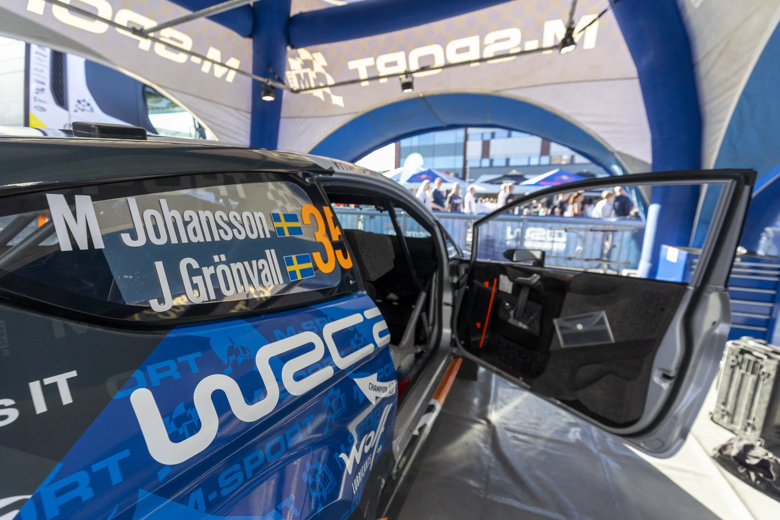 Close-up of a racing car with the names Johansson and Grönvall on the side, parked under a tent at a motorsport event with people and tents in the background.