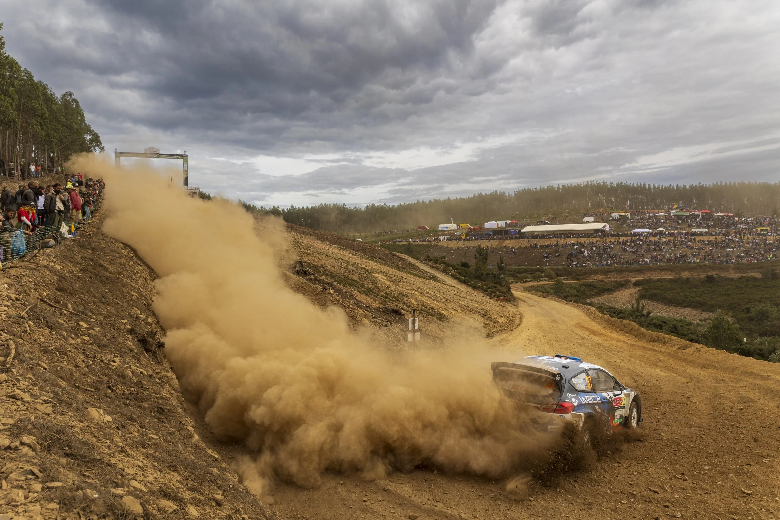 A race car speeding on a dirt track during a rally race, kicking up dust, with spectators watching from the side and a crowd in the background