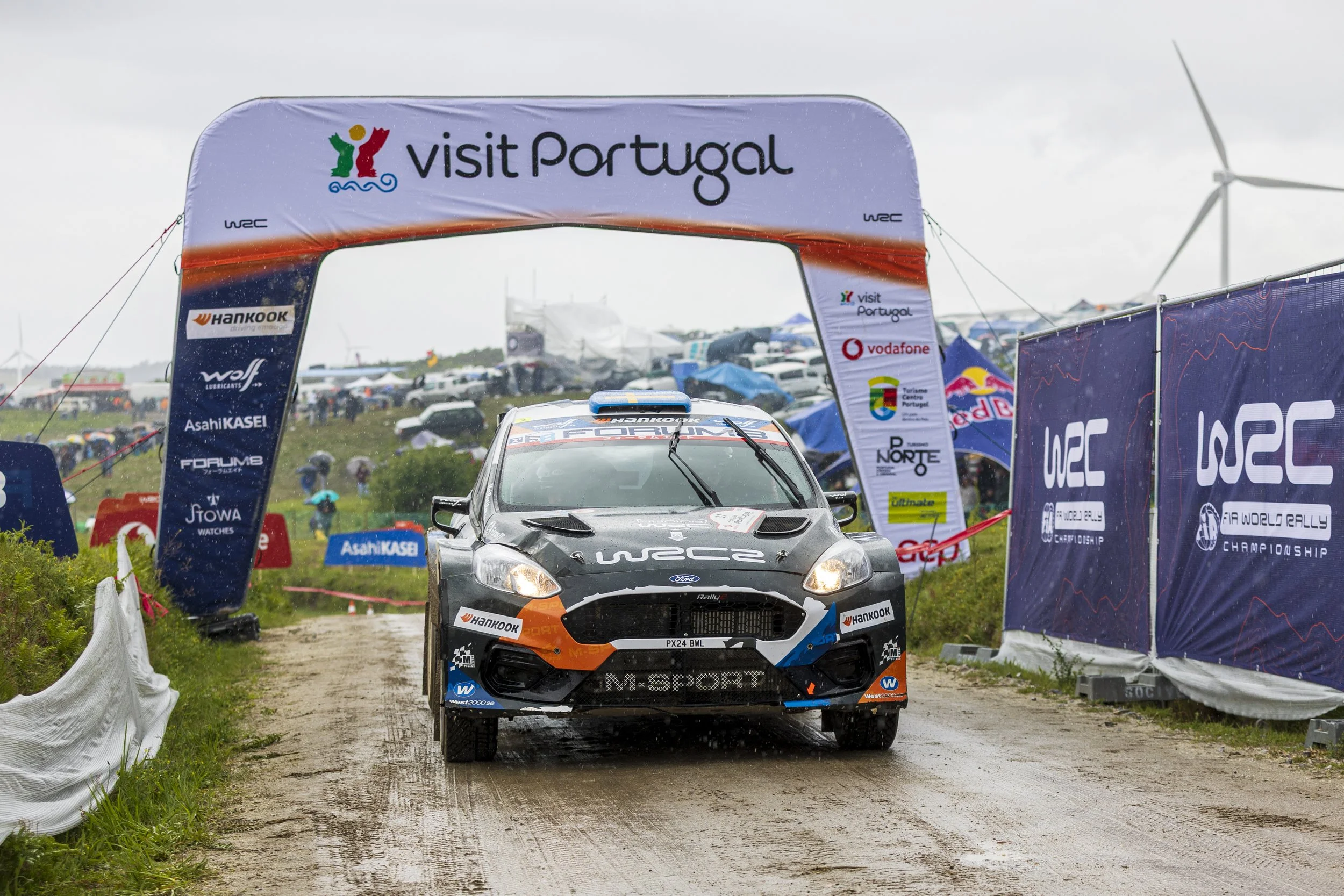A rally car driving on a dirt track at a race event in Portugal, with banners and flags, spectators with umbrellas in the background, and wind turbines in the cloudy sky.