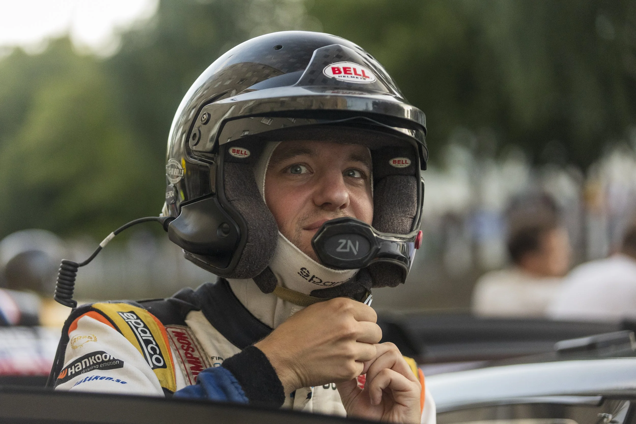 Close-up of a male race car driver wearing a racing helmet and suit, looking confident with a slight smile, in a racing environment.