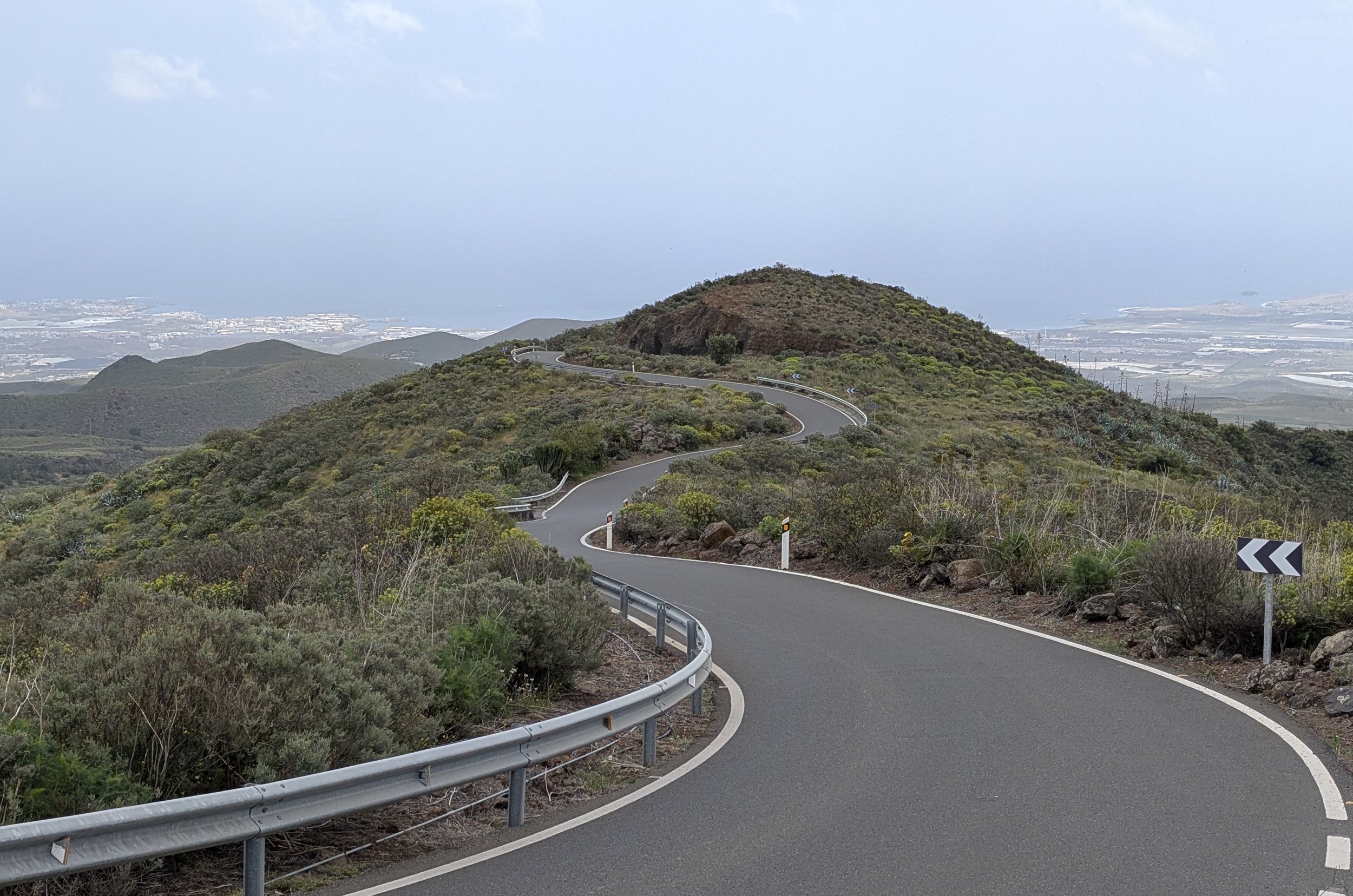 Winding mountain road with guardrails and directional signs, surrounded by green shrubbery and hills under a cloudy sky.