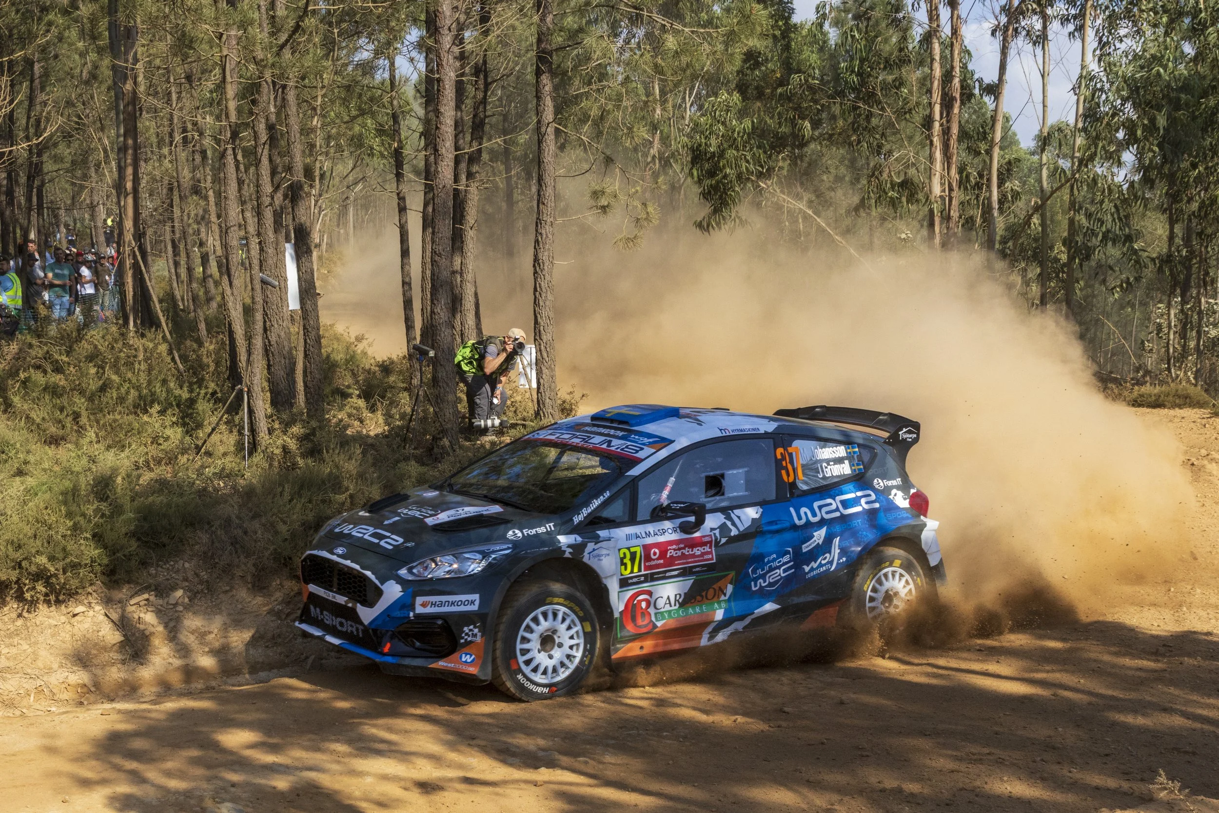 A blue rally car speeding on a dirt road through a forest, kicking up dust, with a crowd watching from the side.