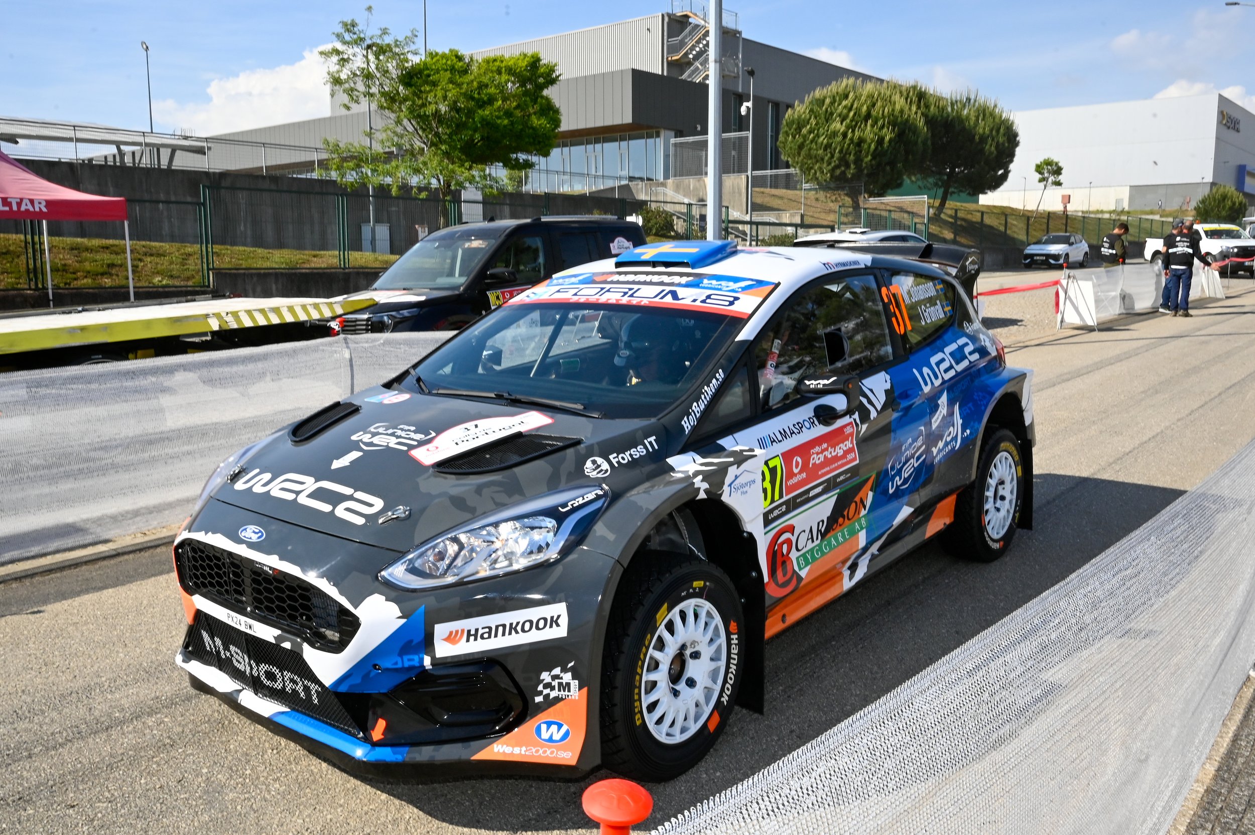 A rally car with sponsorship decals and the number 37 on the side parked on a staging area at a race event. In the background, there is a black truck, several other vehicles, and people preparing near fencing and a building.