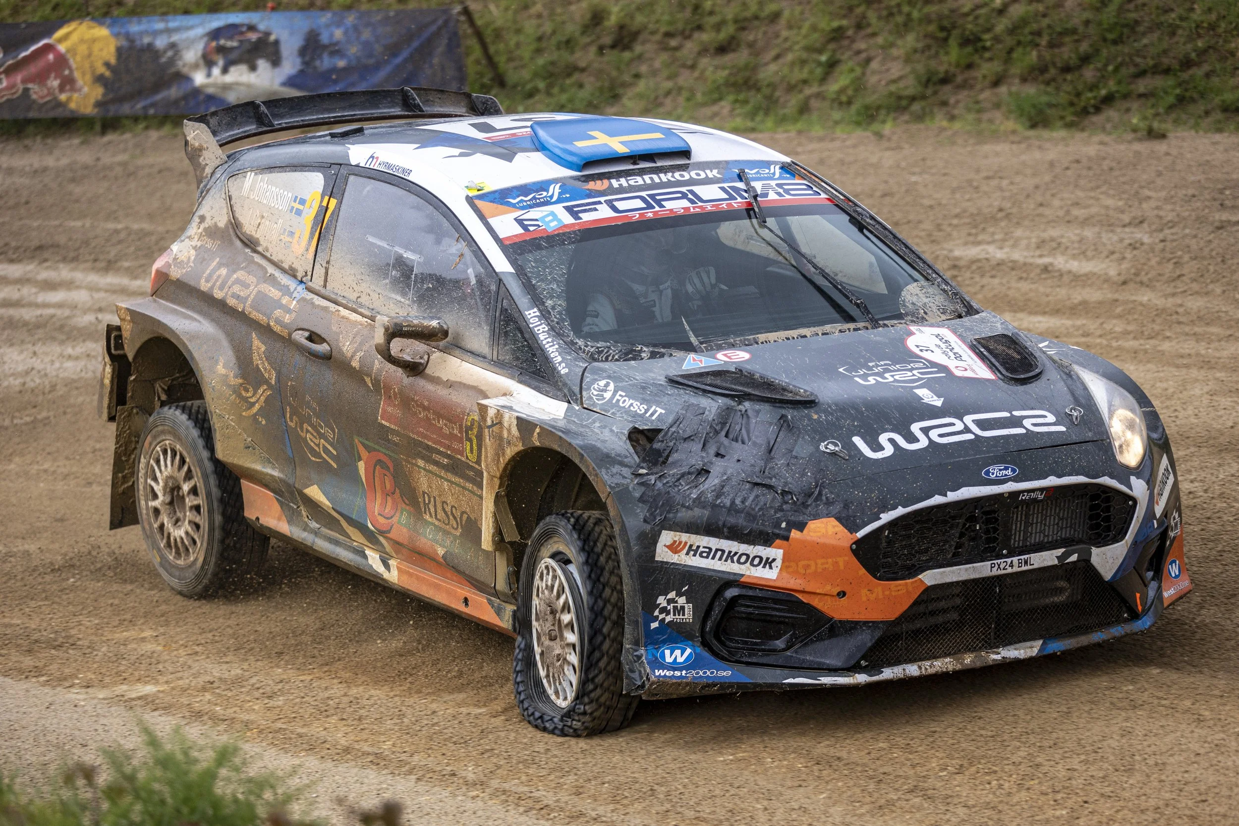 A rally car racing on a dirt track, kicking up dust, with various sponsor logos and decals, including Finnish flags and tire brand logos.