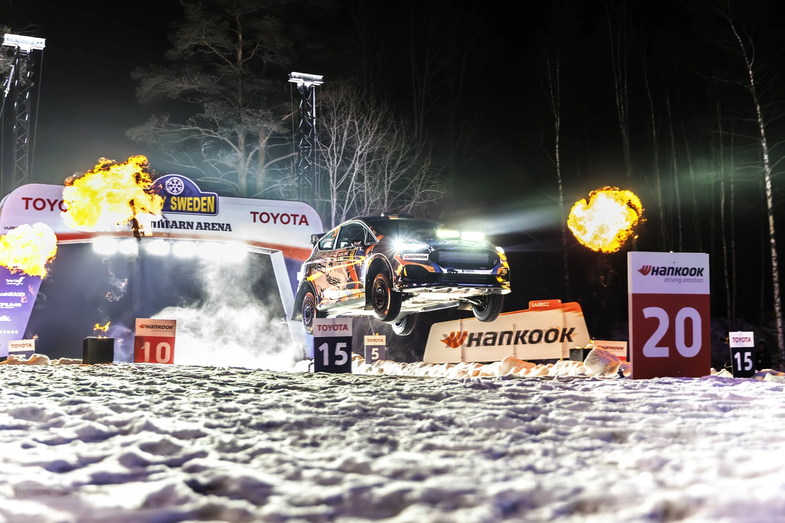 A rally car in mid-air during a nighttime competition with fire and explosion effects, snow-covered ground, and banners displaying sponsor names like Toyota and Hankook.