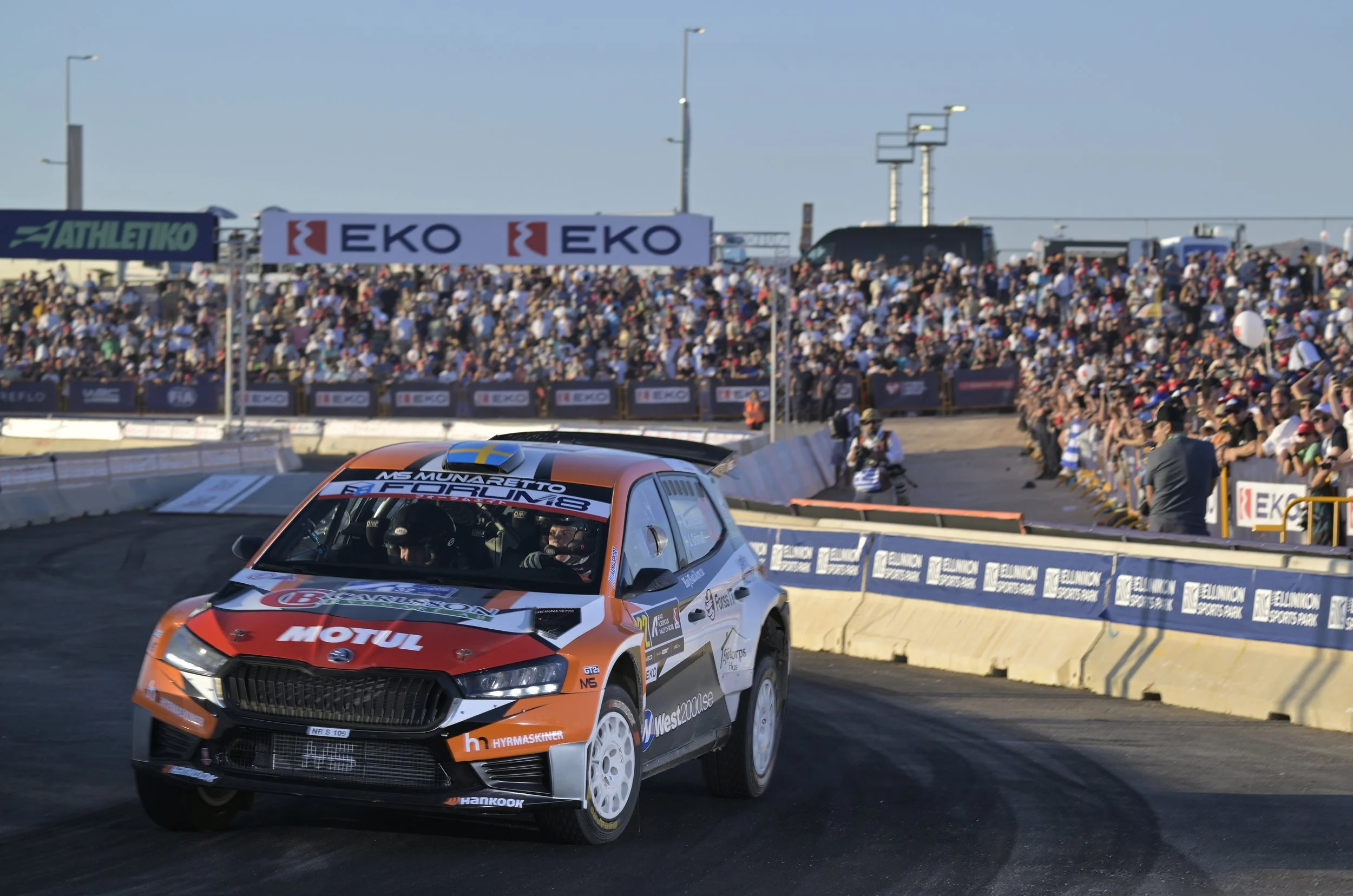 Race car speeding on track with a large crowd of spectators in the background, banners and advertisements visible.