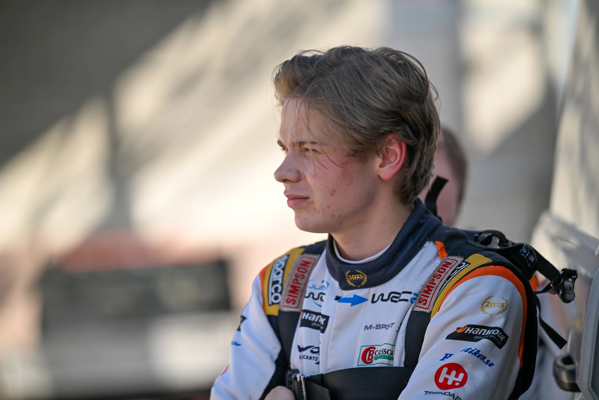 A young race car driver with brown hair and red spots on face, wearing a white racing suit with various sponsor logos, including Hankook, sitting outdoors.