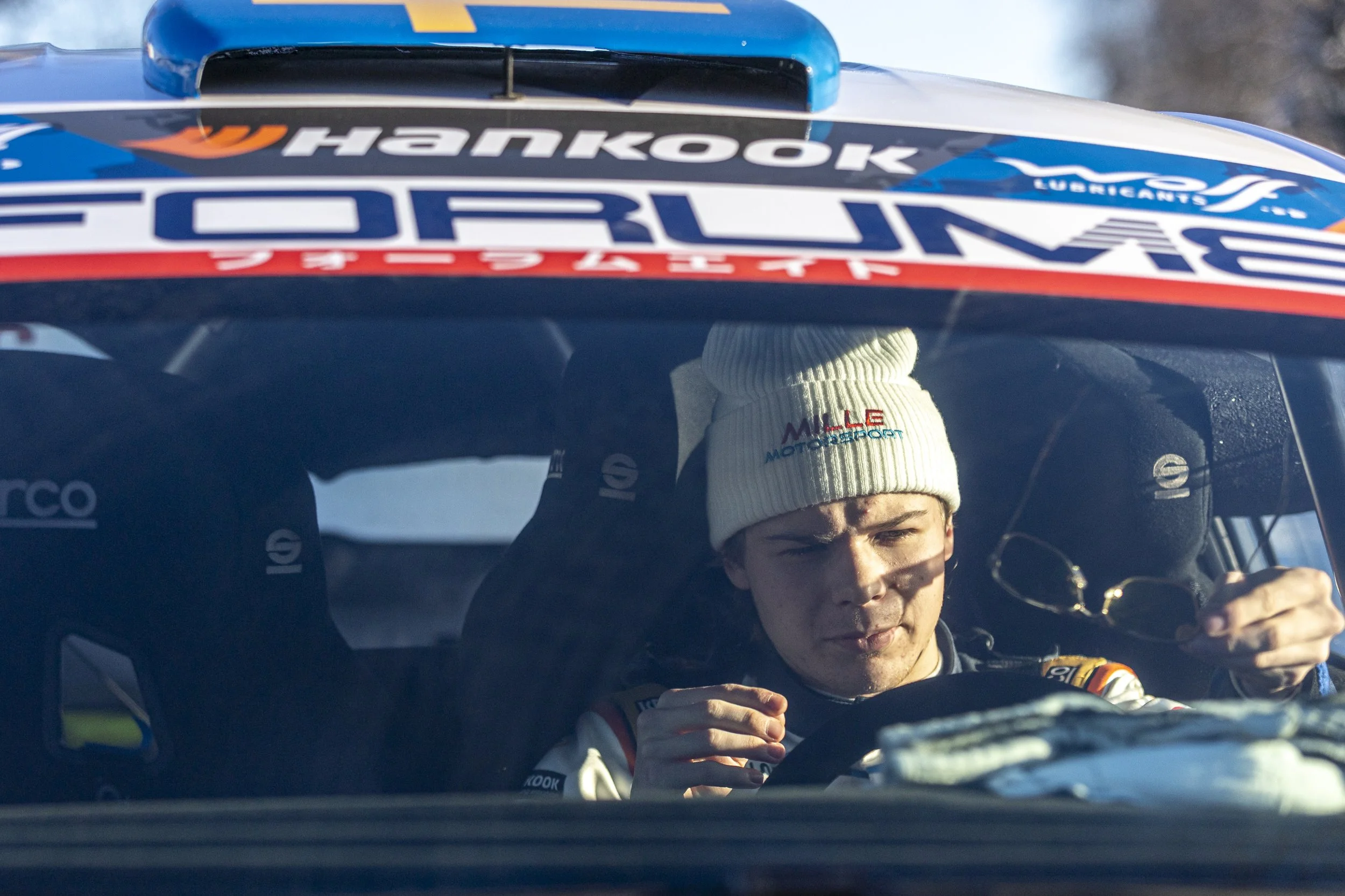 A race car driver inside a race car, wearing a white beanie and racing suit, looking out the windshield. The top of the car's interior and promotional decals are visible.