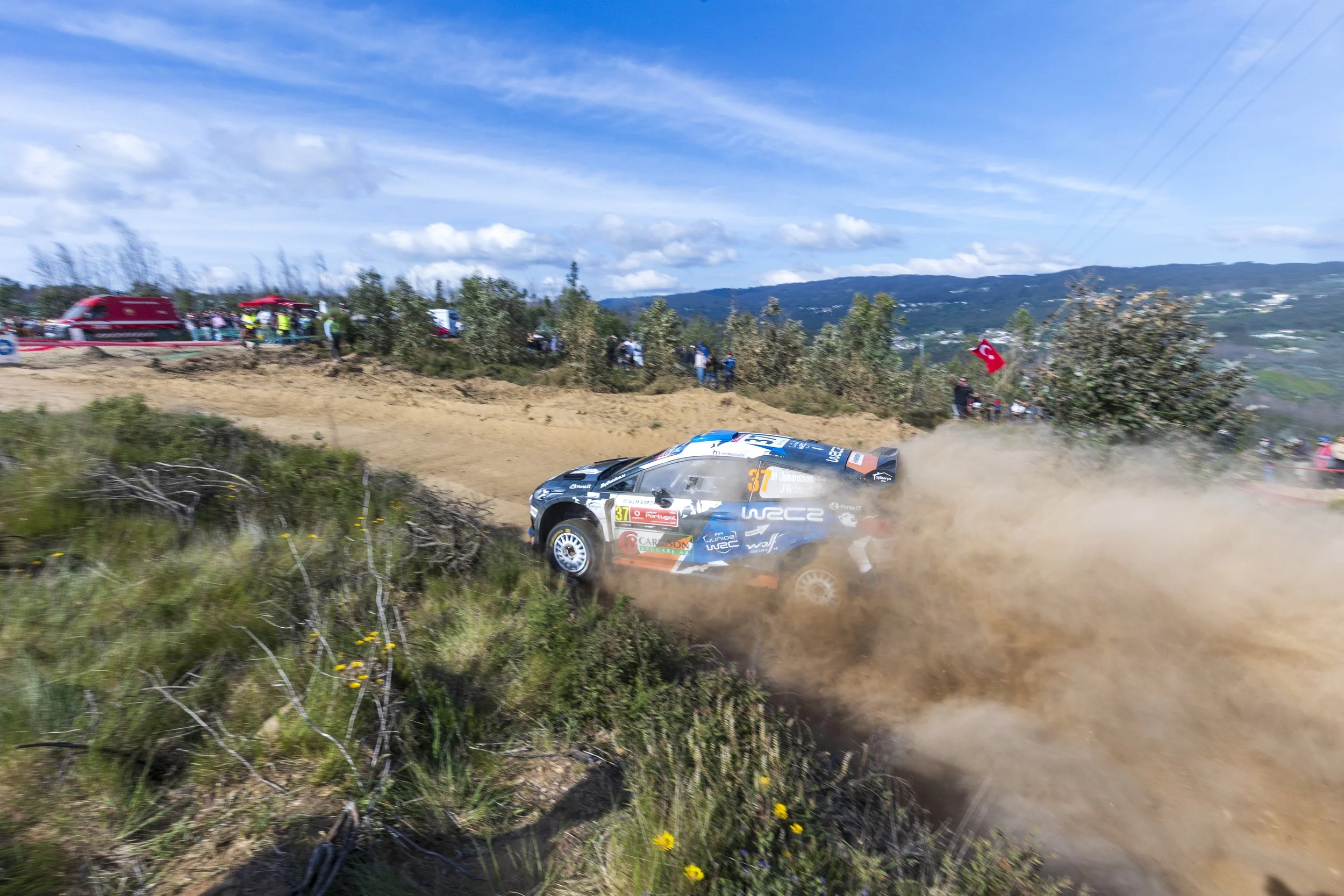 A rally car racing on a dirt track with dust clouds behind it, surrounded by green bushes and spectators in the background, with a blue sky and mountains in the distance.