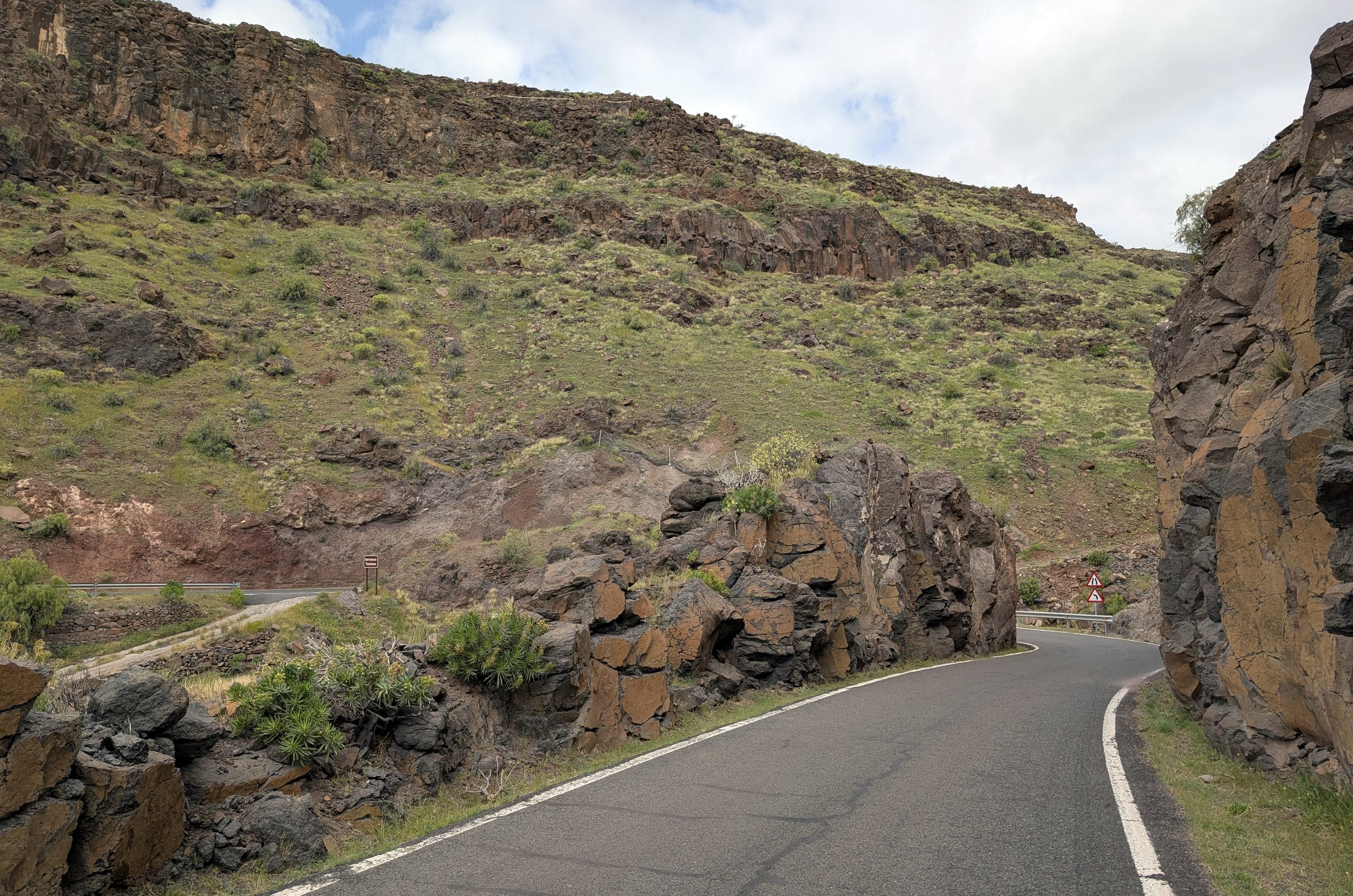 A winding mountain road surrounded by rocky cliffs and green vegetation, with road signs and a small guardrail visible.