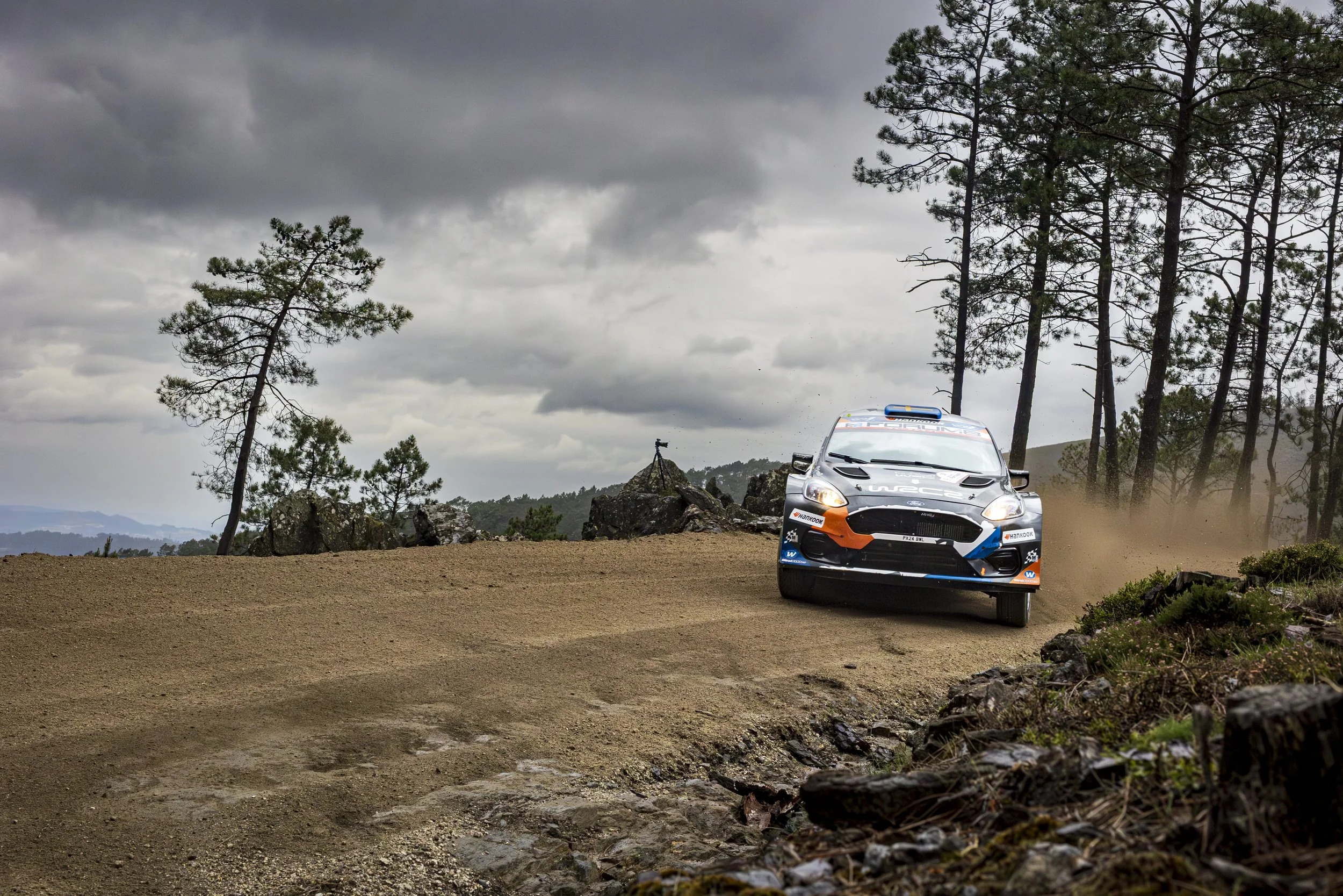 A rally car driving on a dirt road, kicking up dust, surrounded by a forested landscape with tall pine trees and cloudy skies overhead.