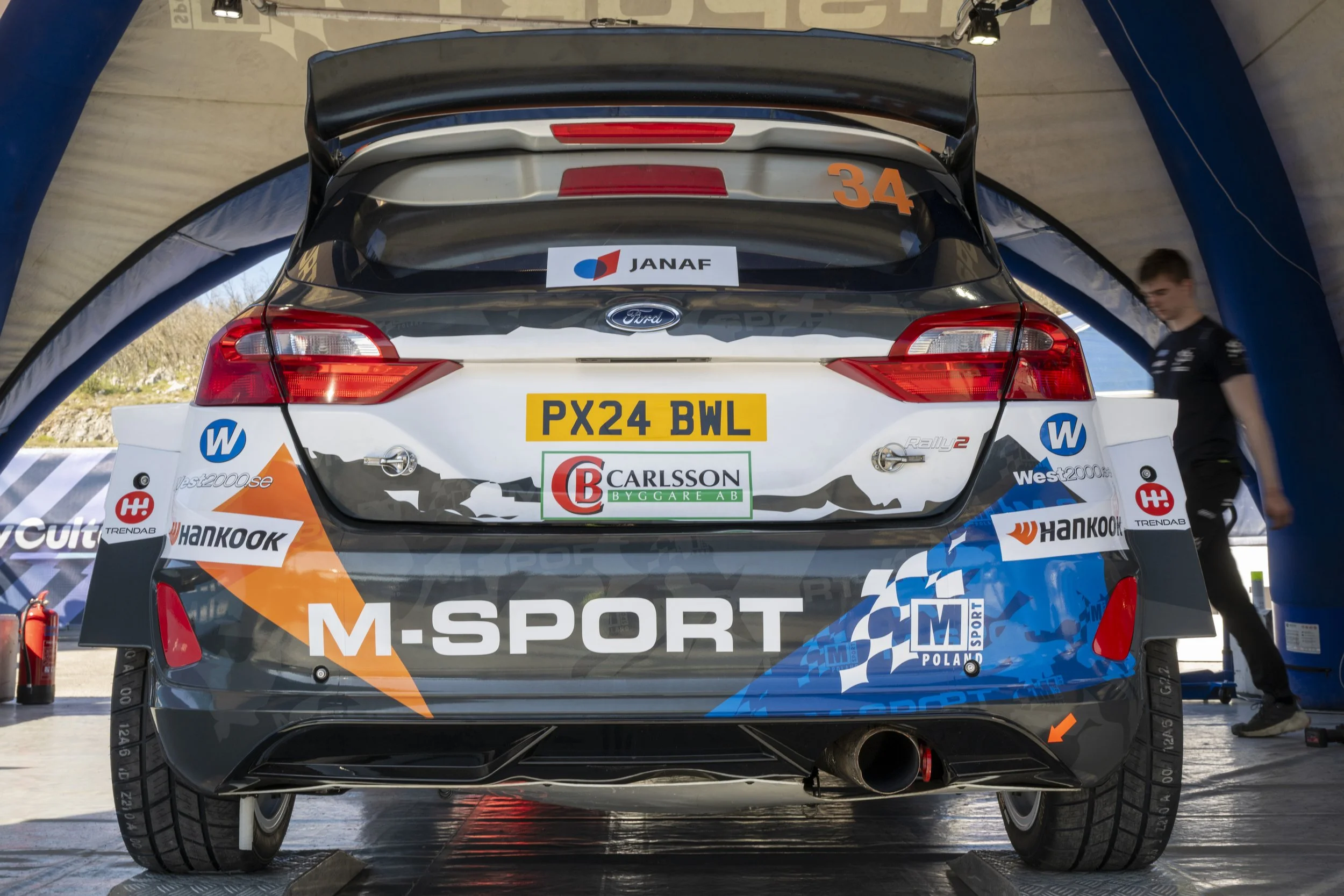Back view of a rally car with sponsor logos, racing number 34, under a tent, with a person on the right side in black shirt.