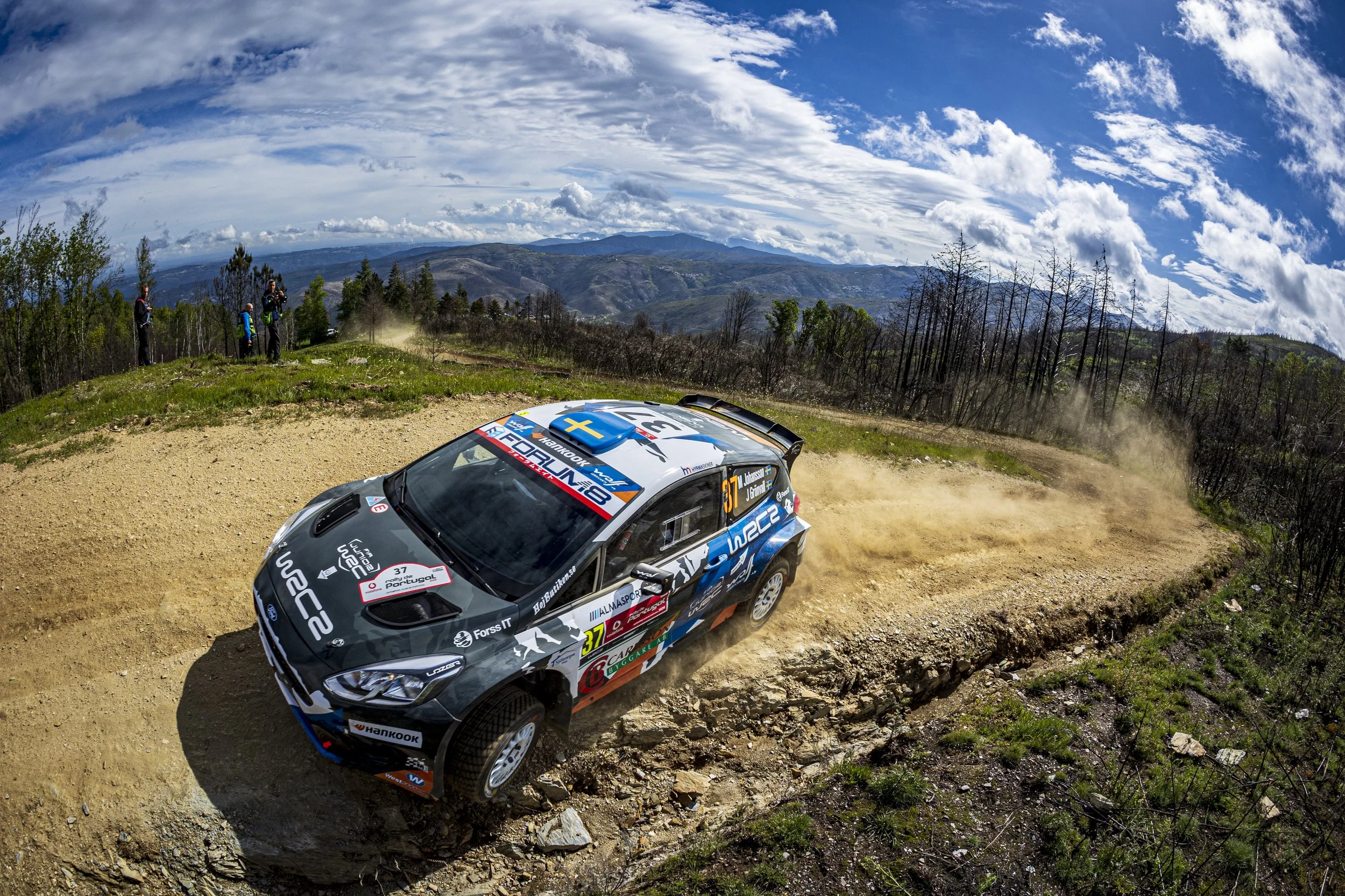 A rally car speeding on a dirt road with dust clouds, surrounded by trees and mountains under a partly cloudy sky.