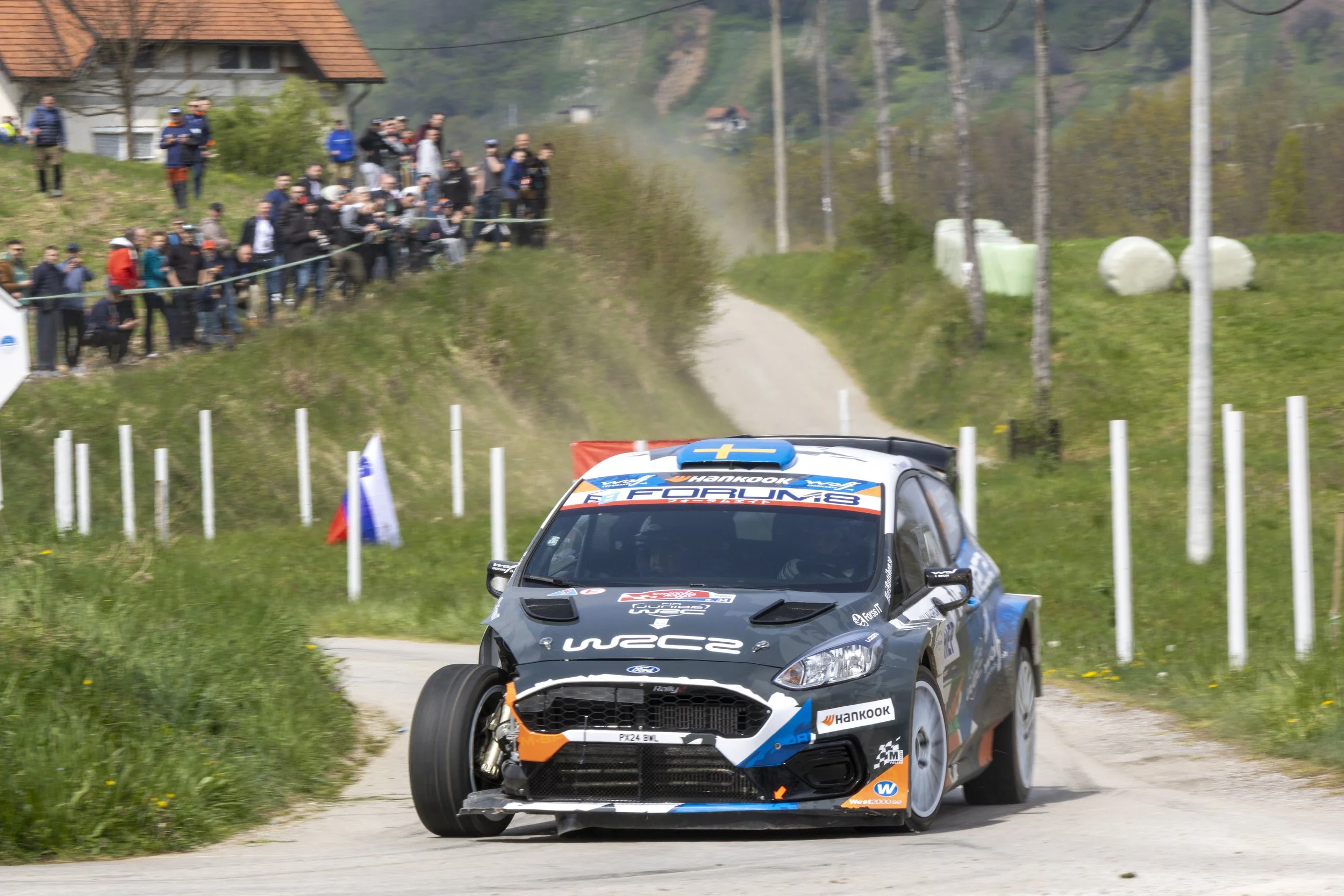 A rally car racing on a dirt track, with a crowd of spectators on a hillside and trees in the background.