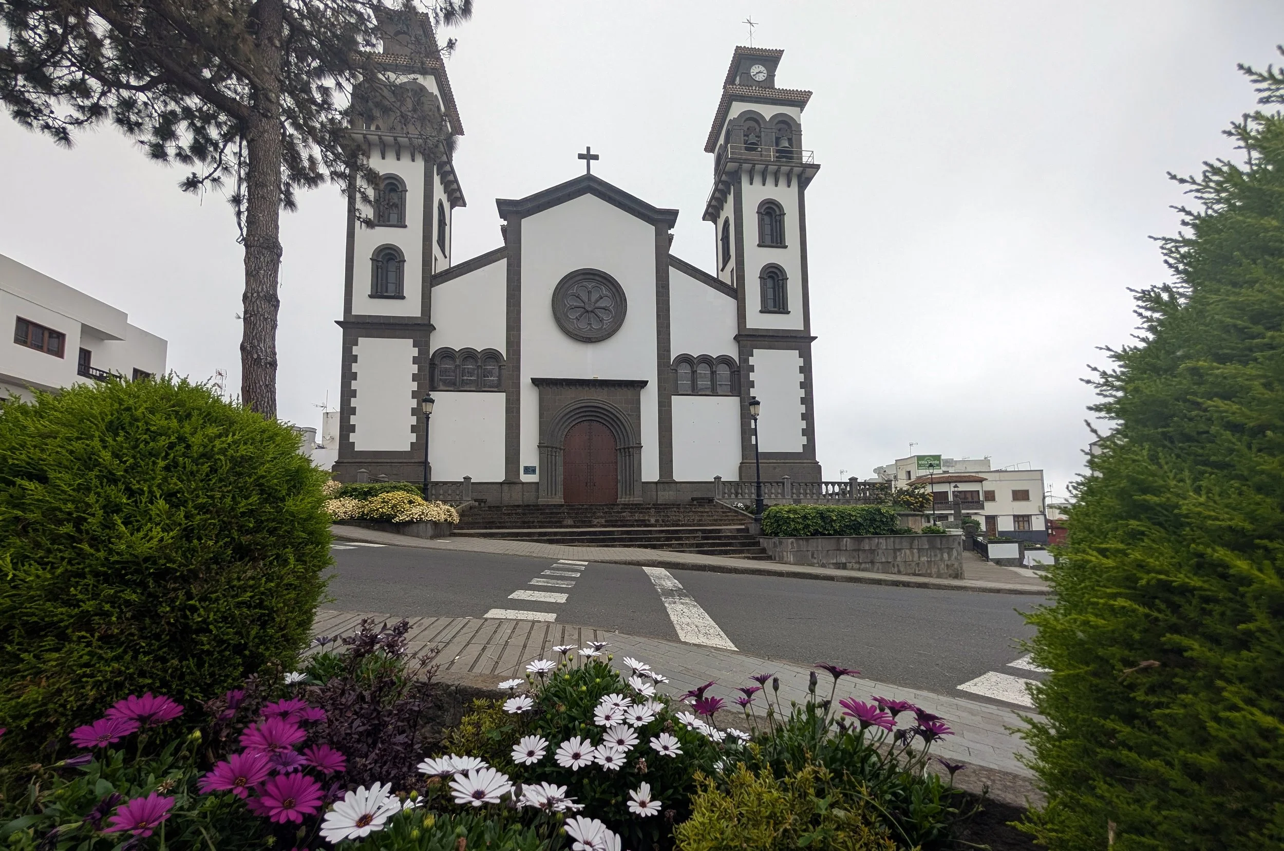 A church with two tall towers, a central façade with a rose window, and a cross on top, surrounded by greenery and colorful flowers in the foreground.