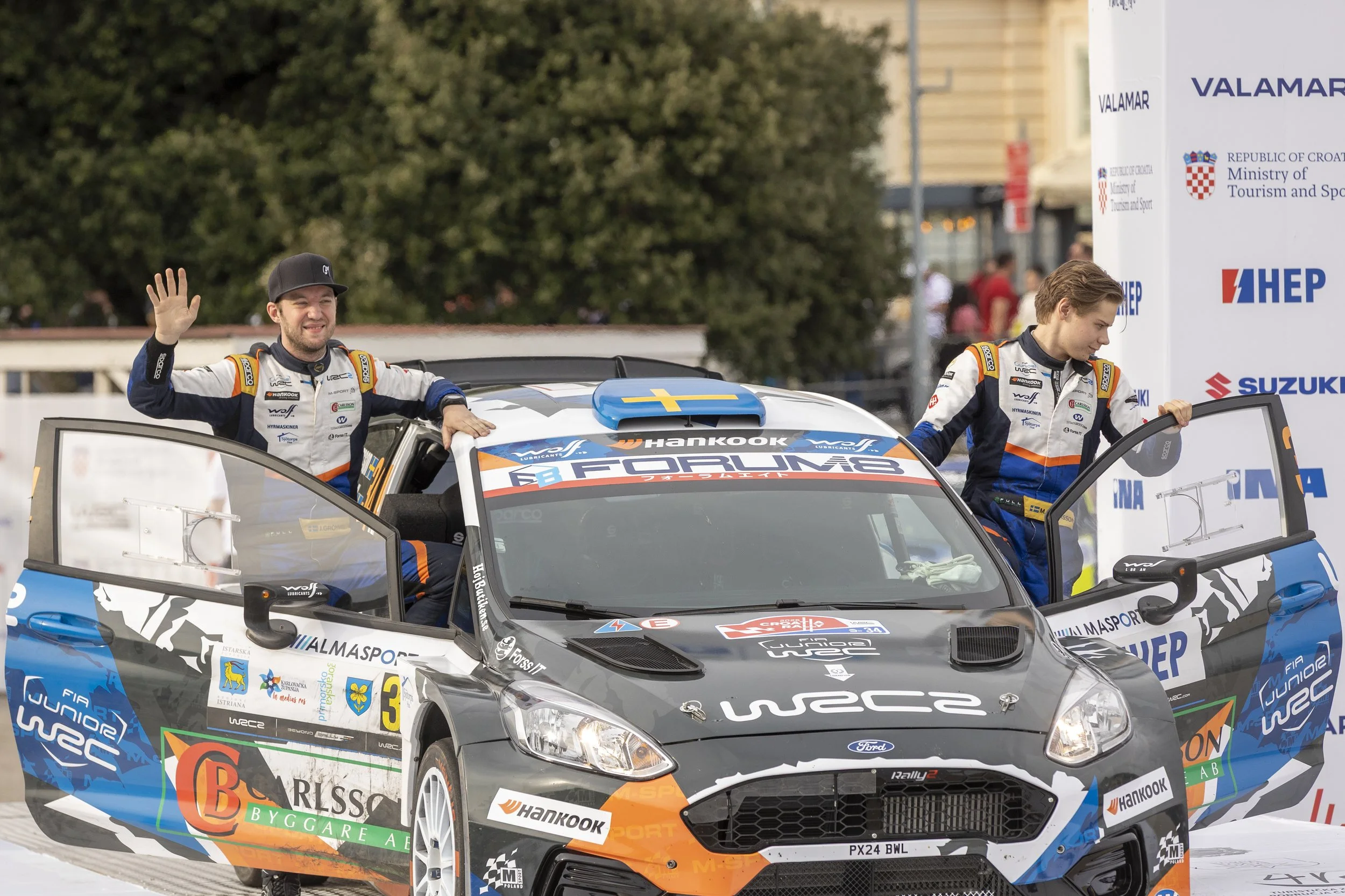 Two race car drivers in racing suits standing in and outside a rally car, smiling and waving. The scene appears to be at a rally car event with sponsorship banners in the background.