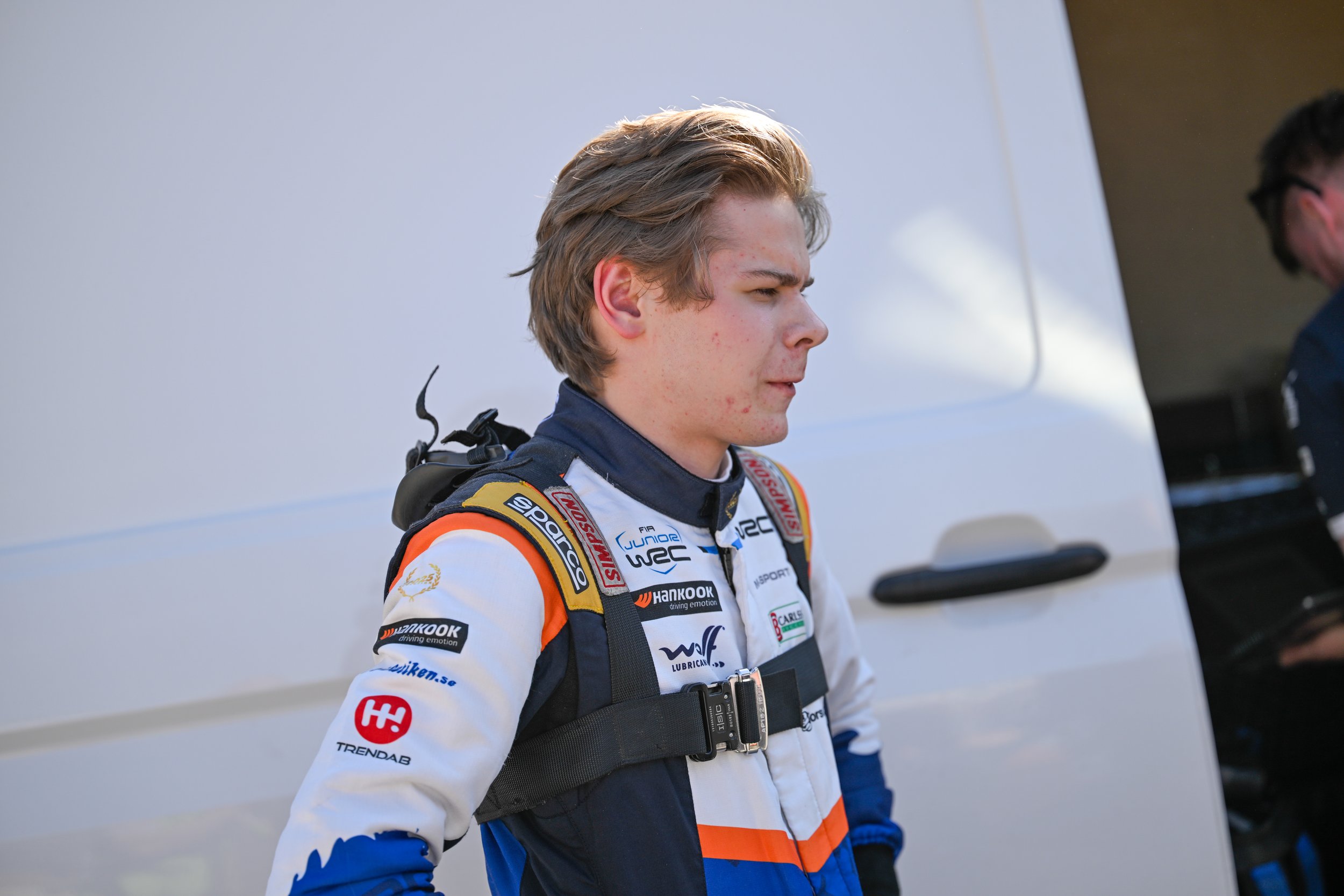 Young male race car driver wearing a racing suit with sponsor logos, standing outdoors near a white trailer or truck.