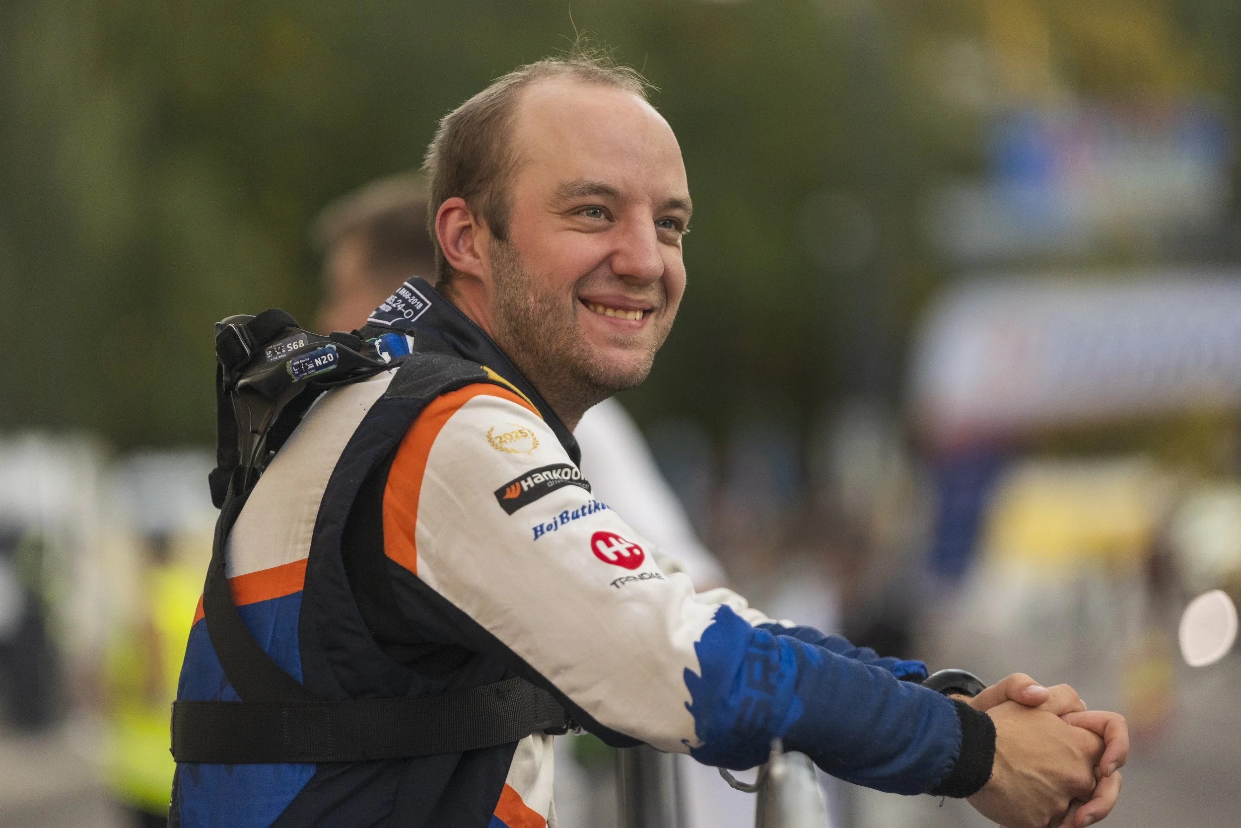A smiling man in a racing suit with sponsor logos, standing outdoors with a blurred background.