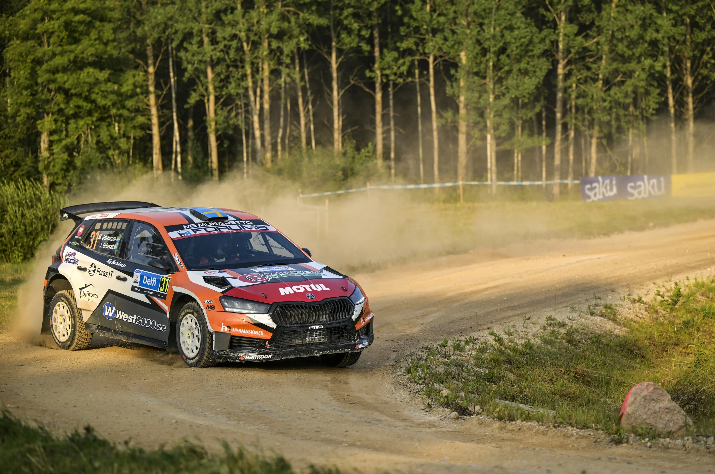 A rally car with sponsor logos driving on a dirt road surrounded by trees and dust.