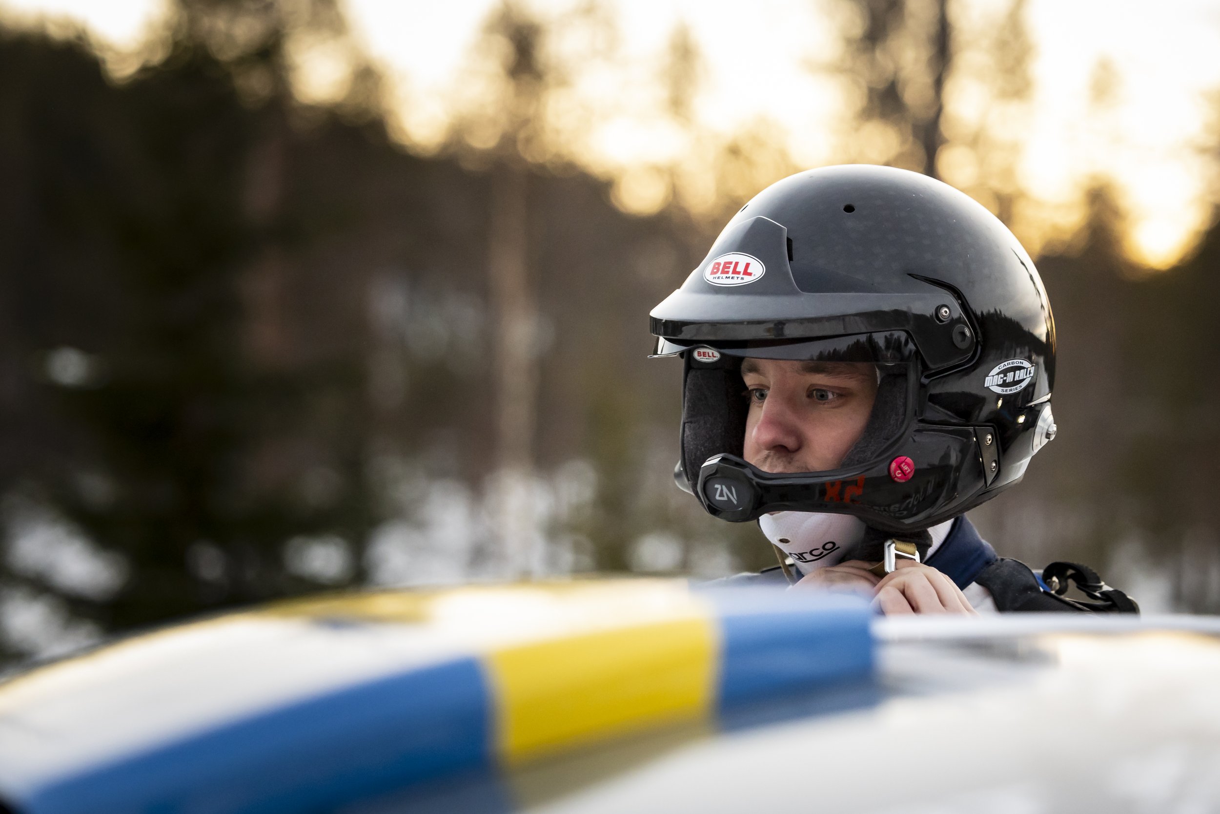A person wearing a racing helmet looking out from a vehicle, with a blurred background of trees and sunset.