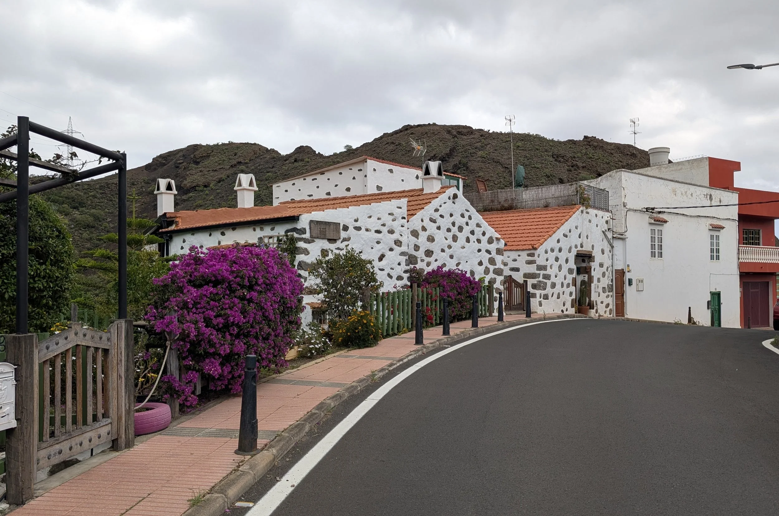A street with a curved black asphalt road and a pink sidewalk border, lined with black bollards. There are white houses with red-tiled roofs, blooming pink bougainvillaea, and scenic mountains in the background under cloudy skies.