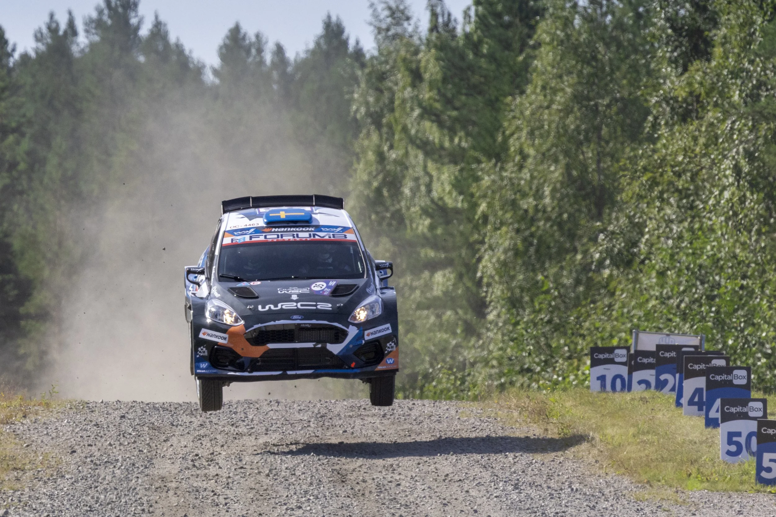 A rally car in mid-air jumping over a gravel road during a race, surrounded by green trees and sponsor signs along the road.