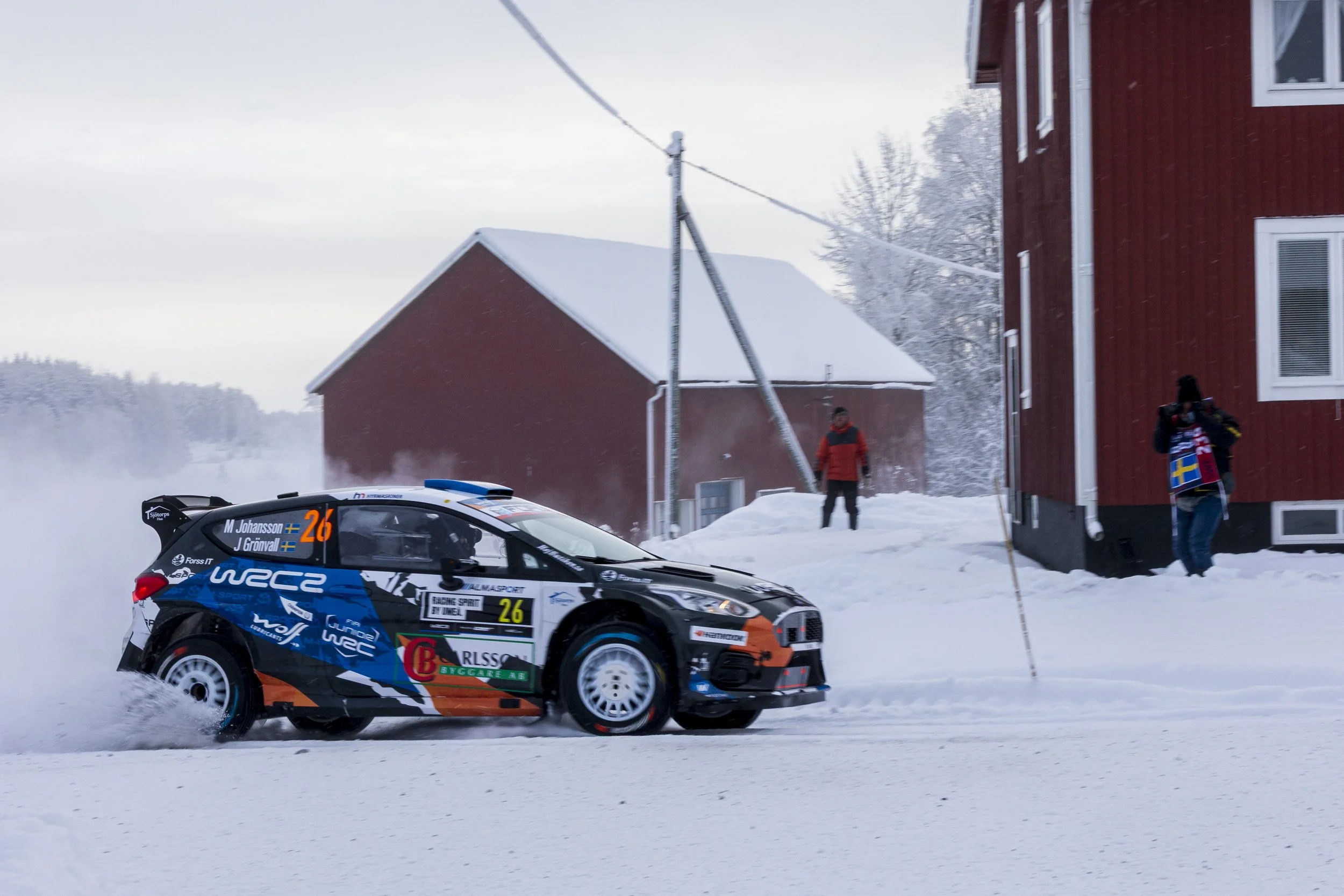 A rally car driven by M. Johansson and J. Granvall speeds through a snowy landscape, kicking up snow behind it during a winter rally event, with red buildings and snow-covered trees in the background.